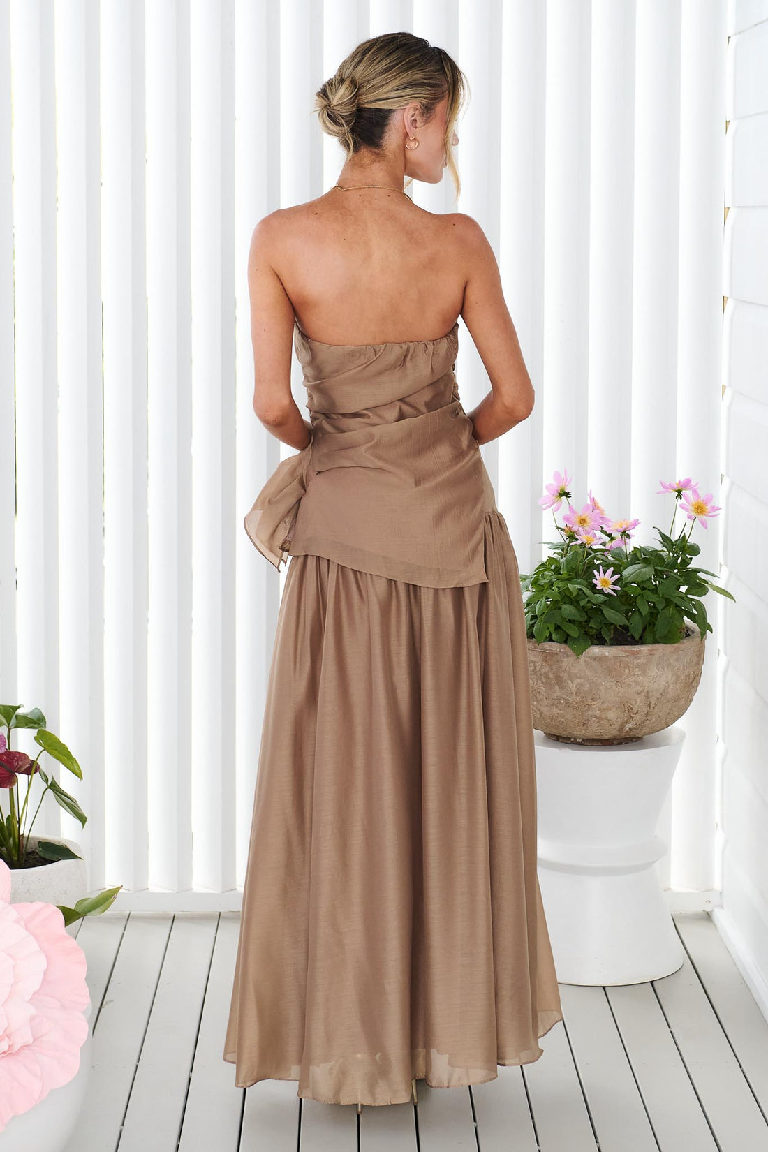 Back view of a woman wearing the minnie + MAXXIE strapless mocha brown event set with a ruched bodice and flowing maxi skirt, standing in front of a white panelled wall.