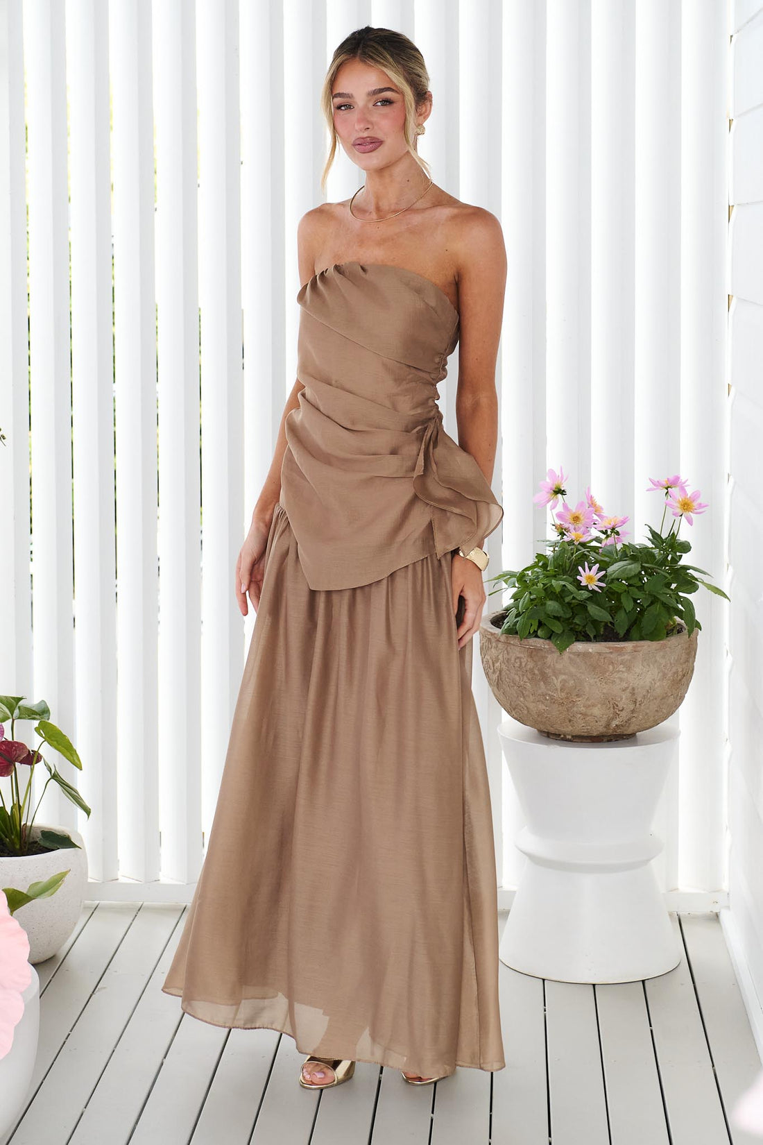 A woman wearing the minnie + MAXXIE strapless mocha brown event set with a ruched bodice and flowing maxi skirt, standing in front of a white panelled wall.