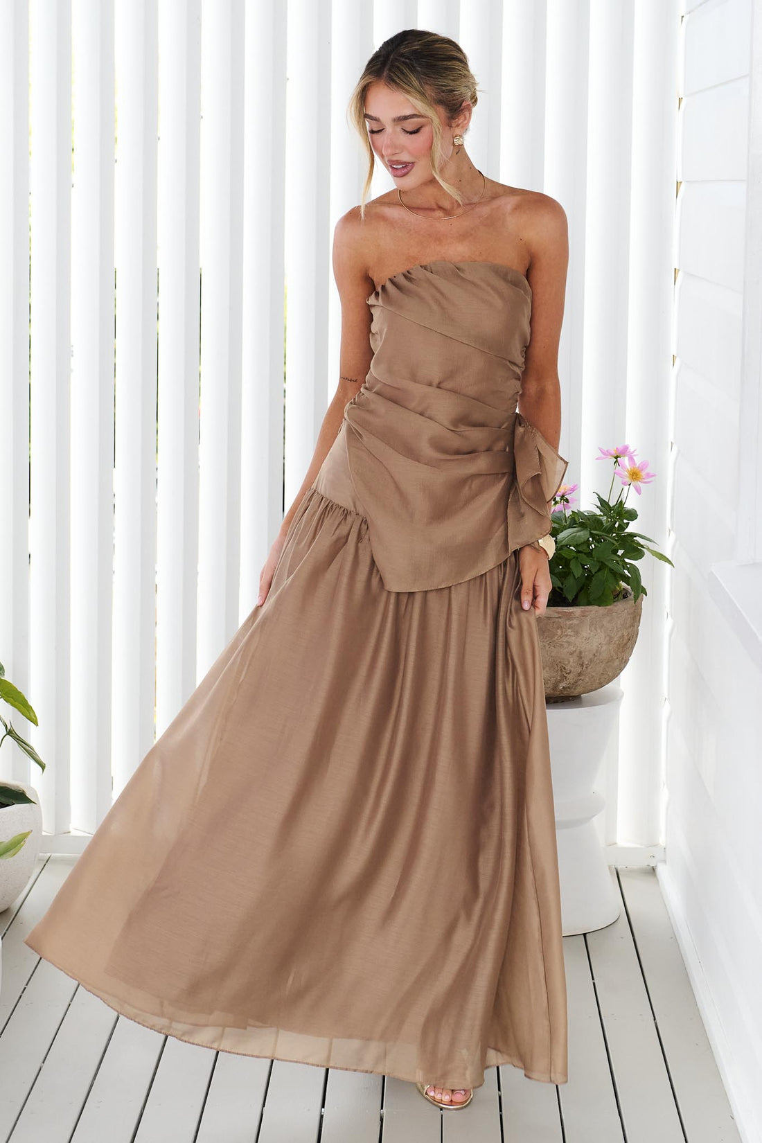 A woman wearing the minnie + MAXXIE strapless mocha brown event set with a ruched bodice and flowing maxi skirt, standing in front of a white panelled wall.