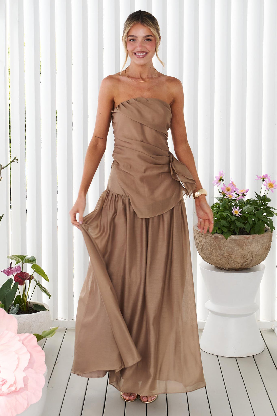 A woman wearing the minnie + MAXXIE strapless mocha brown event set with a ruched bodice and flowing maxi skirt, standing in front of a white panelled wall.