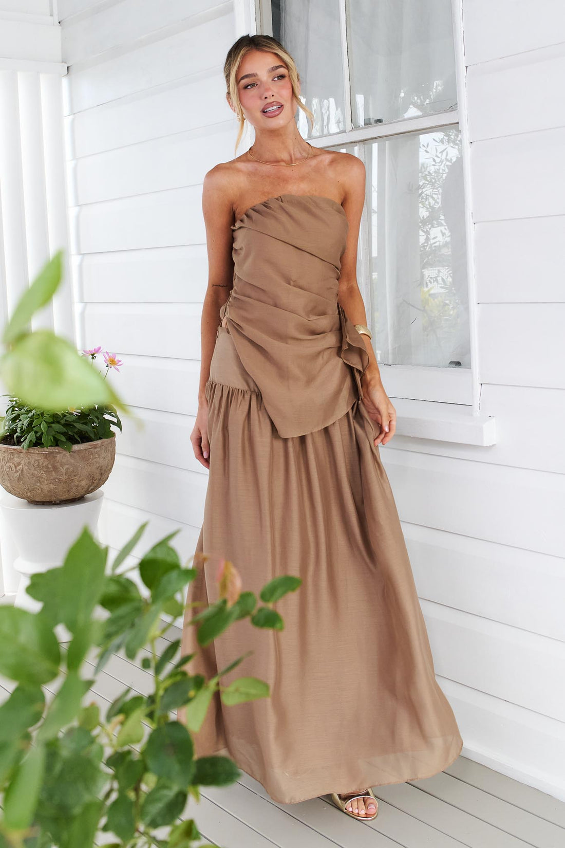 A woman wearing the minnie + MAXXIE strapless mocha brown event set with a ruched bodice and flowing maxi skirt, standing in front of a white window.
