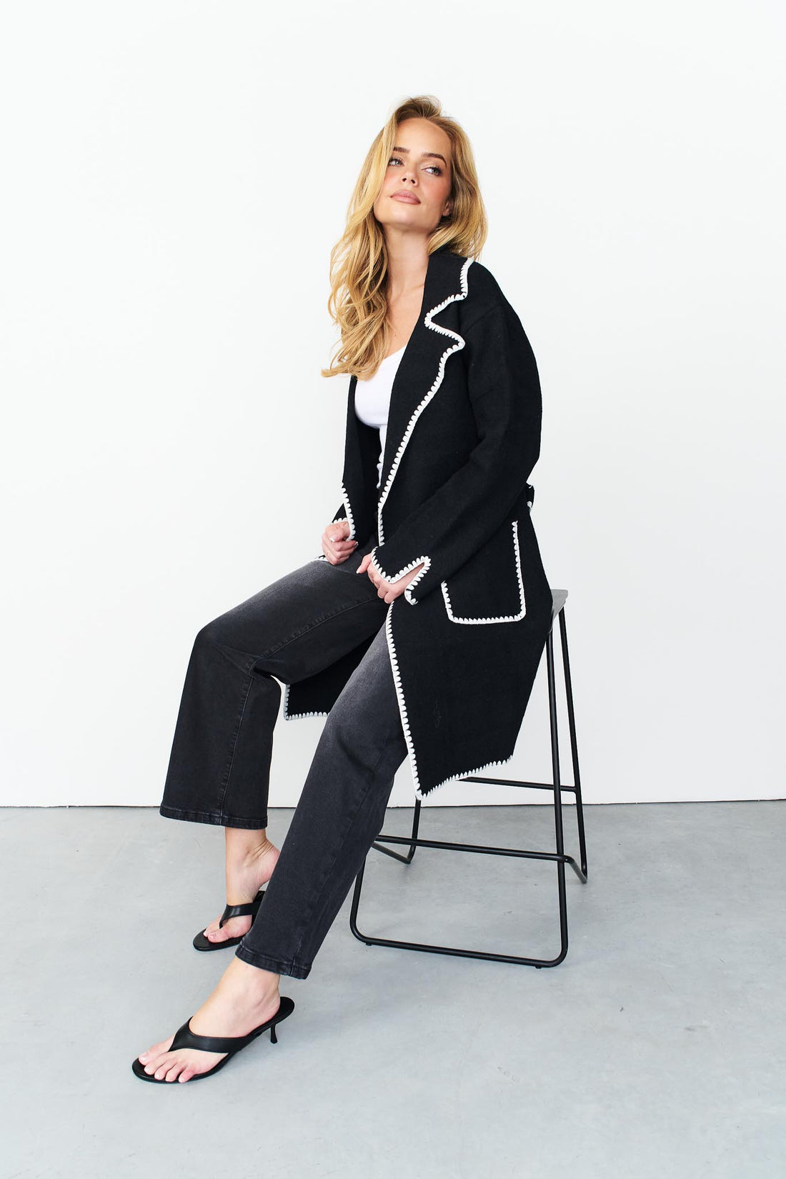 Woman sitting on a stool wearing minnie + MAXXIE wool blend black Kinsey jacket with washed black denim jeans.