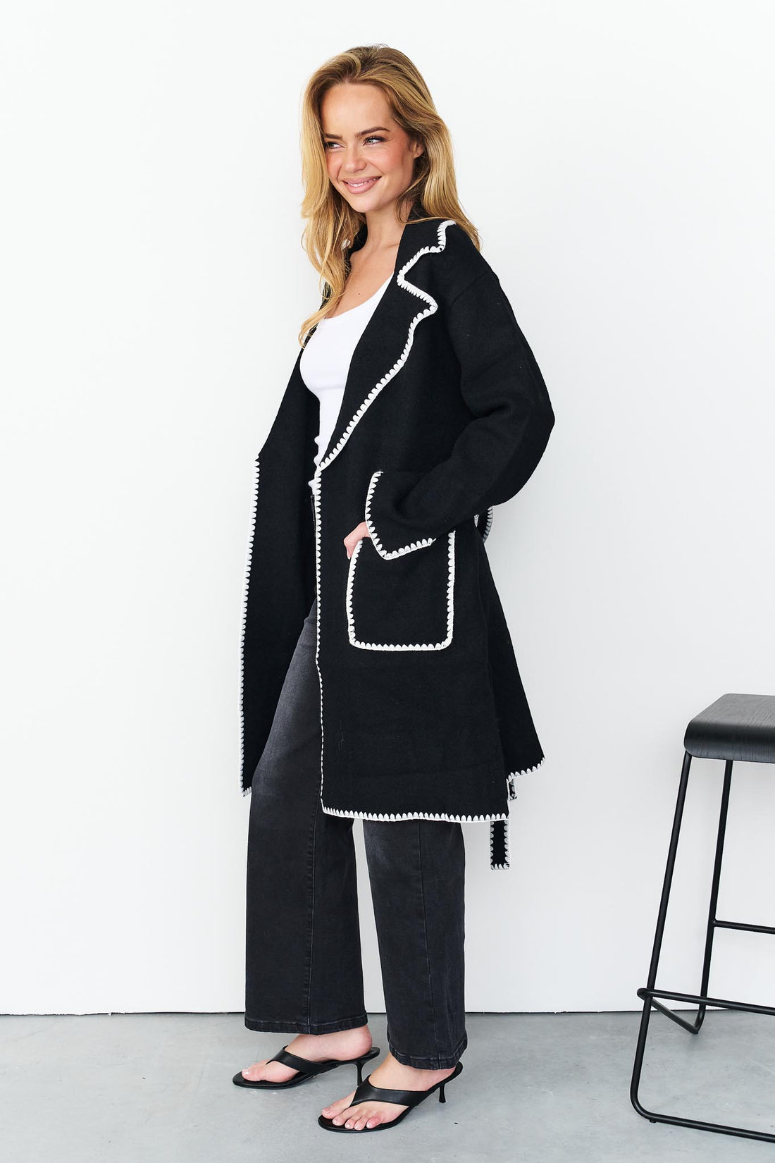 Side view of woman wearing minnie + MAXXIE wool blend black Kinsey jacket with white blanket stitch trim, a white top, and washed black denim jeans.