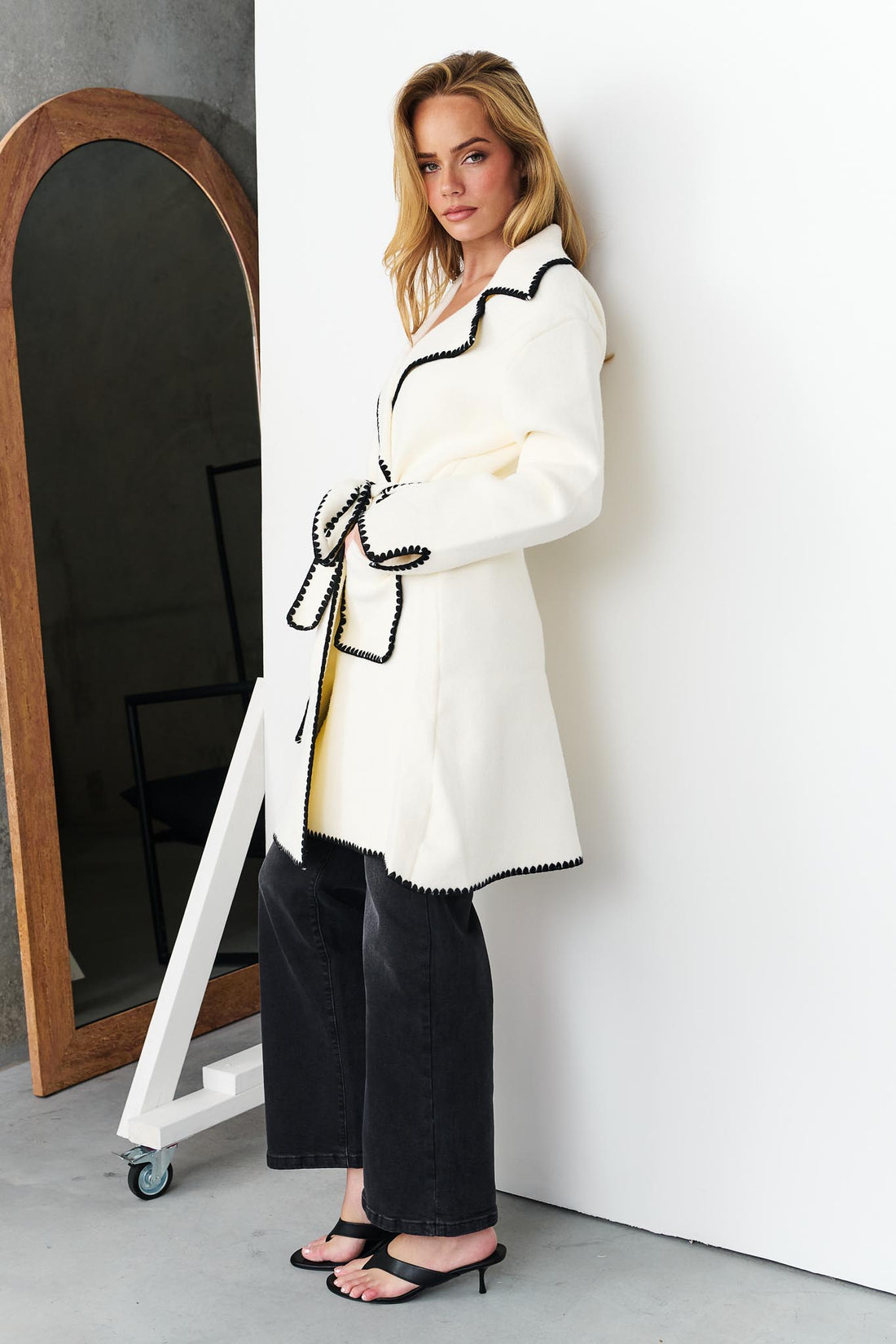 Woman wearing minnie + MAXXIE wool blend ivory Kinsey jacket with black blanket stitch trim, standing next to a full length mirror.