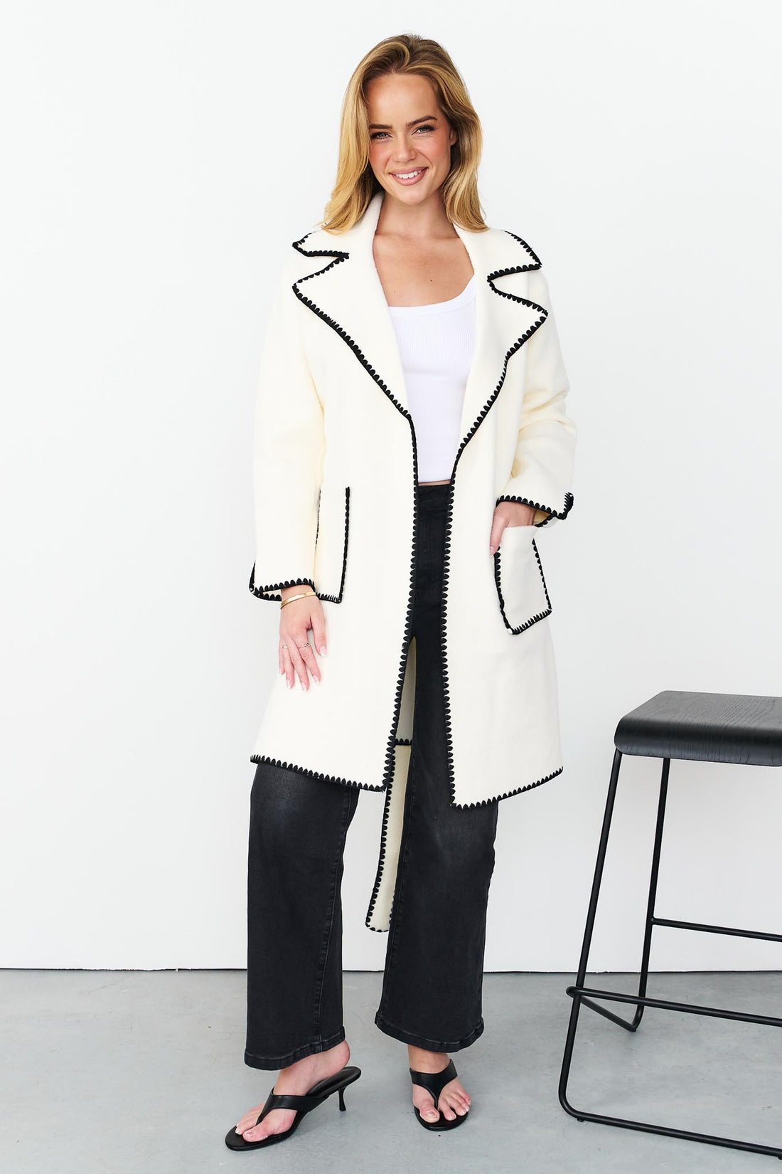 Woman wearing minnie + MAXXIE wool blend ivory Kinsey jacket with black blanket stitch trim, white top and washed black denim jeans.