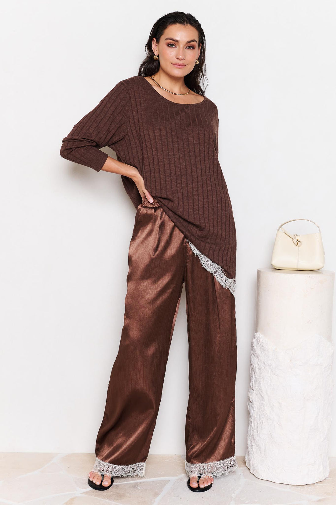 Woman wearing a brown ribbed knit top and satin pants with lace trim, standing against a white background.