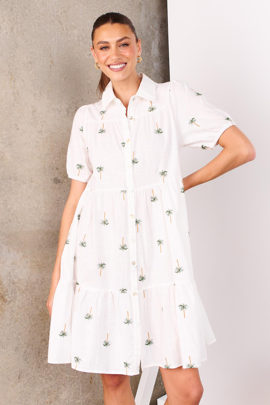 A woman wearing the minnie + MAXXIE Laguna white A-line shirt dress with short-sleeves, a collared neckline and palm embroidery, standing in front of a white and concrete wall.