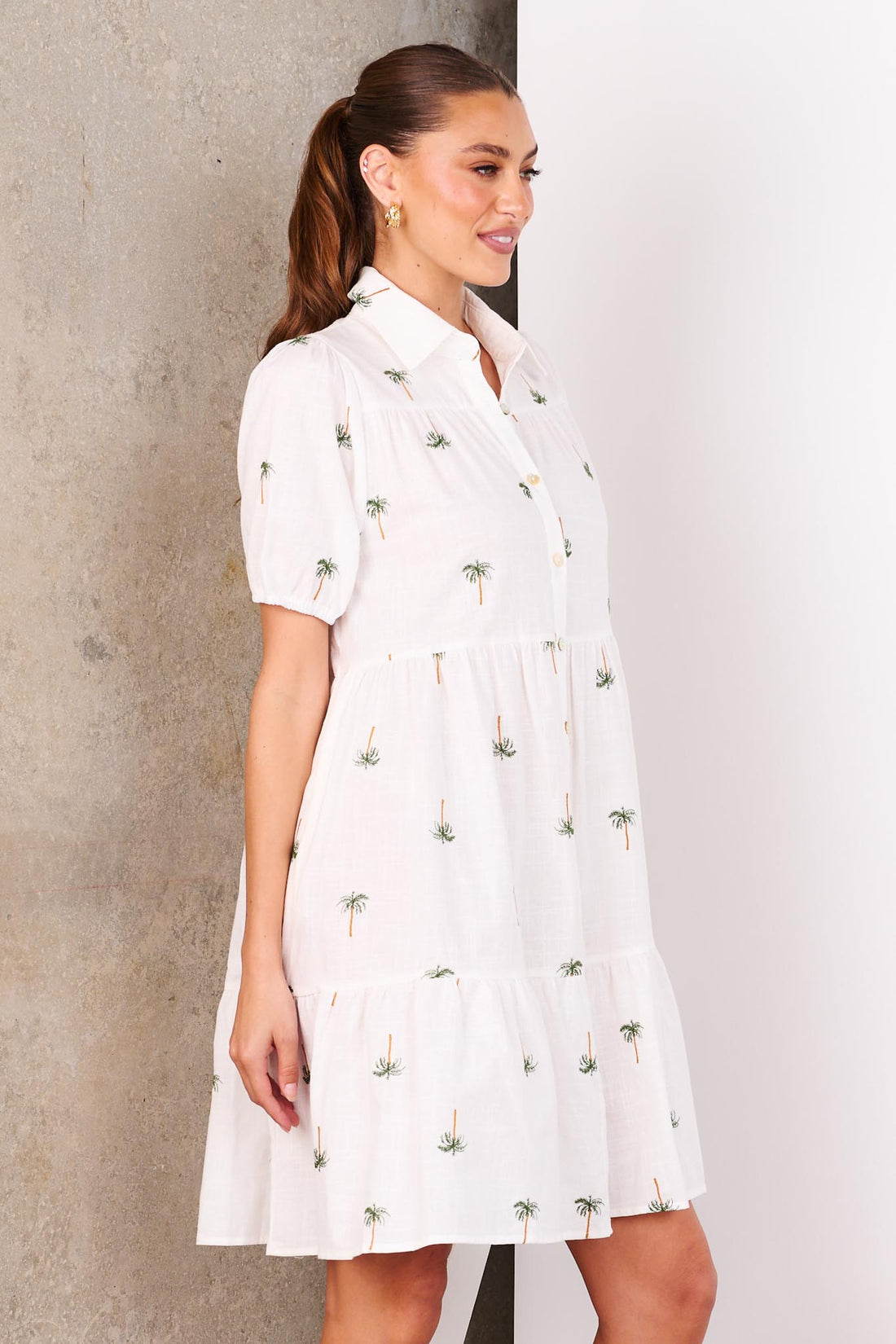 A woman wearing the minnie + MAXXIE Laguna white A-line shirt dress with short-sleeves, a collared neckline and palm embroidery, standing in front of a white and concrete wall.