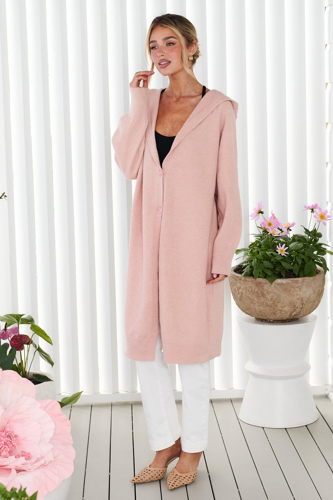 A woman wearing the minnie + MAXXIE long, button front Lennox cardigan in a soft blush pink, worn over a black top and white jeans, standing in front of a white panelled wall.