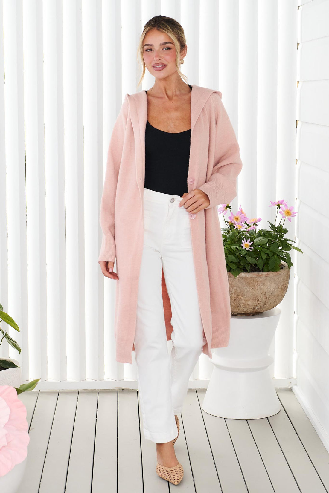 A woman wearing the minnie + MAXXIE long, open-front Lennox cardigan in a soft blush pink, worn over a black top and white jeans, standing in front of a white panelled wall.
