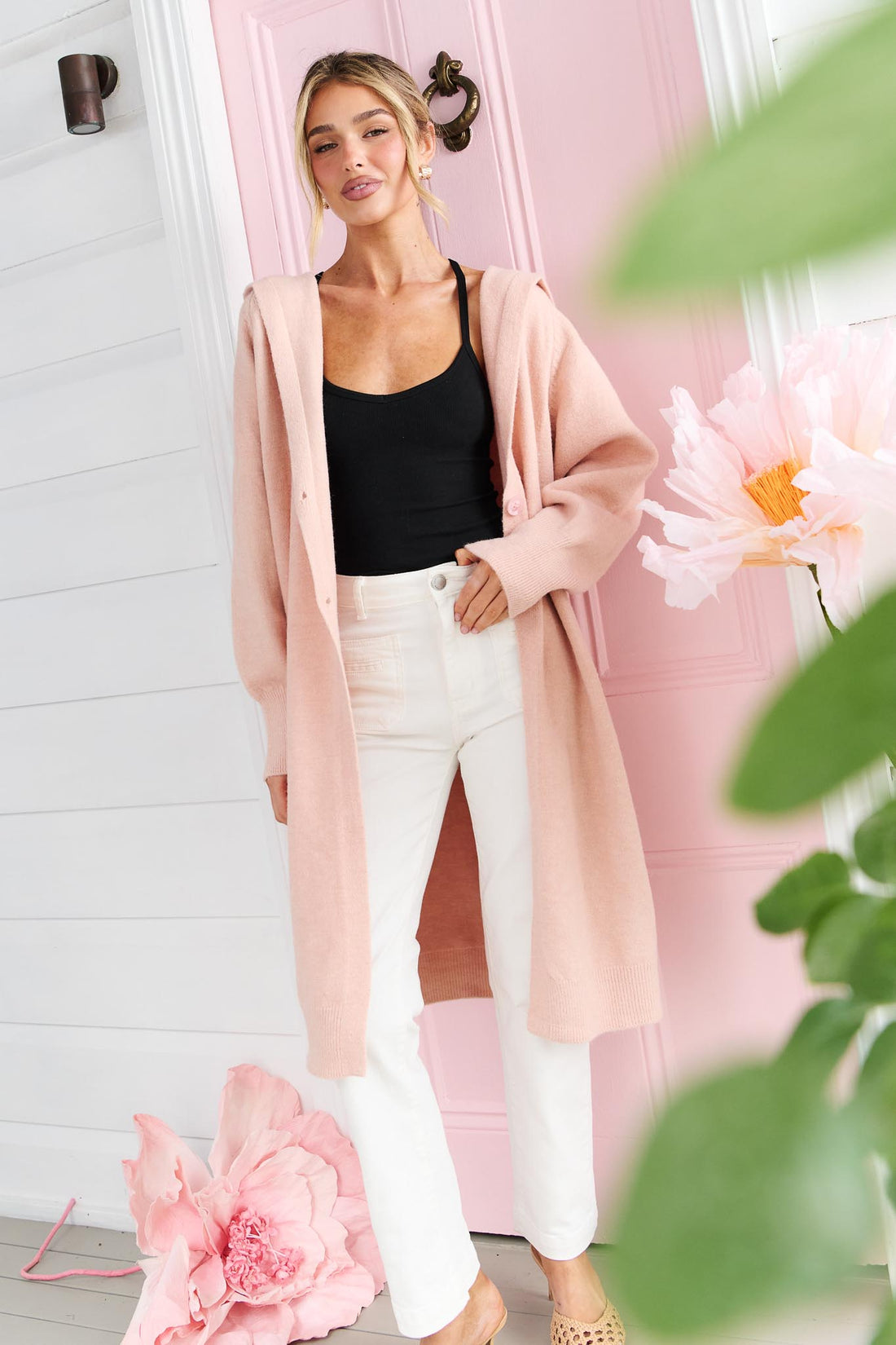 A woman wearing the minnie + MAXXIE long, open-front Lennox cardigan in a soft blush pink, worn over a black top and white jeans, standing in front of a pink door with decorative pink flowers.