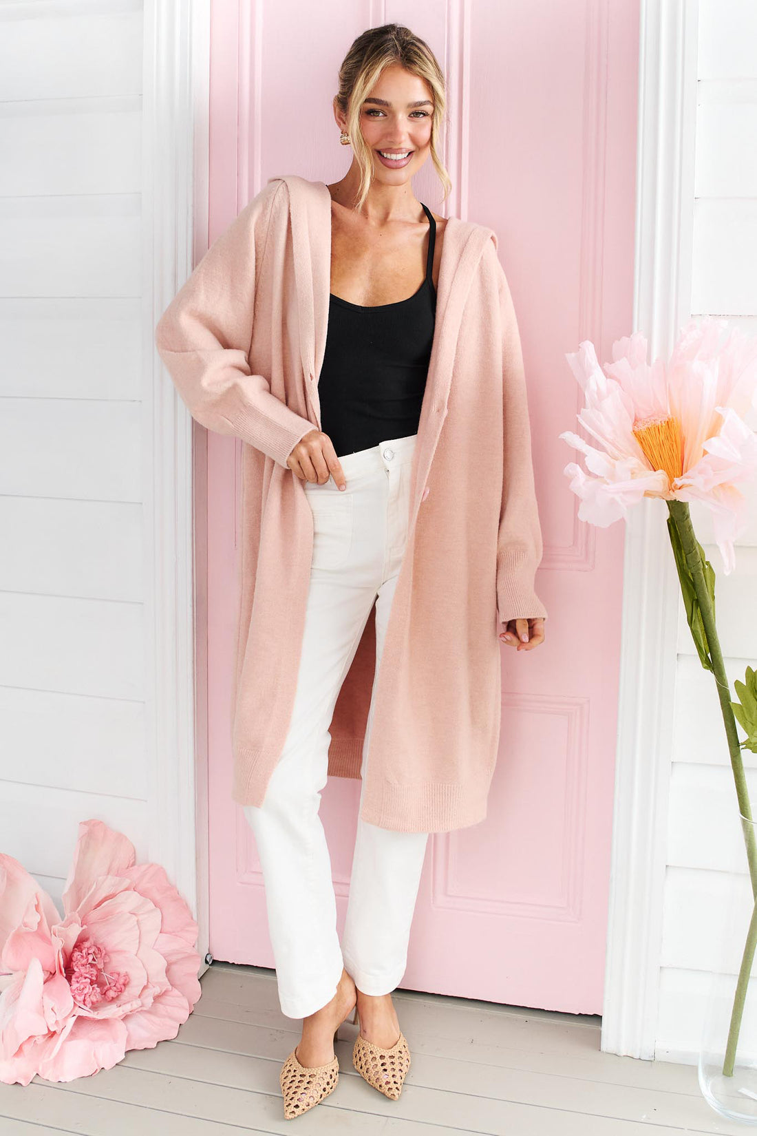 A woman wearing the minnie + MAXXIE long, open-front Lennox cardigan in a soft blush pink, worn over a black top and white jeans, standing in front of a pink door with decorative pink flowers.