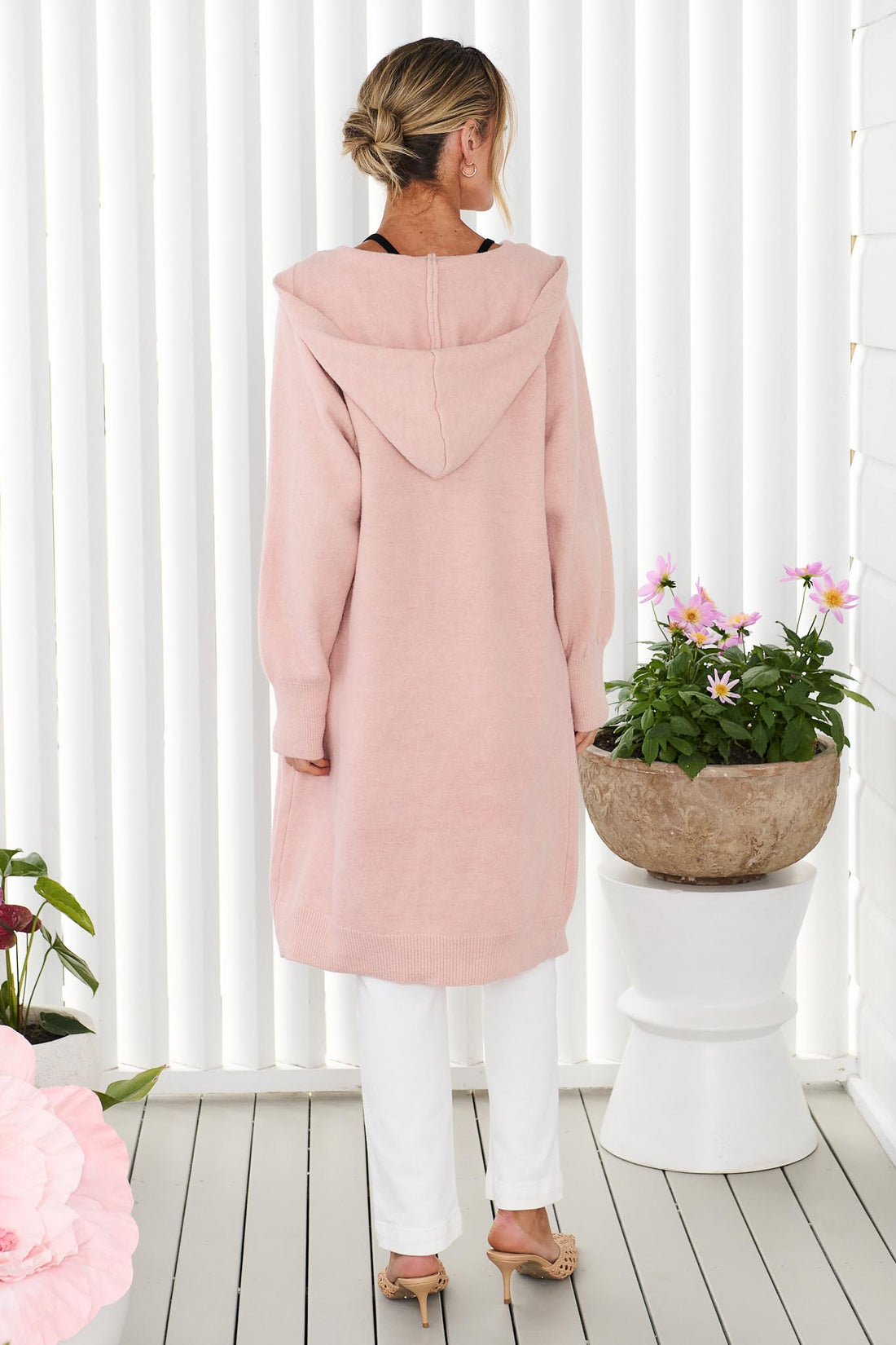 Back view of a woman wearing the minnie + MAXXIE long Lennox cardigan in soft blush pink, worn over white jeans, standing in front of a white panelled wall.