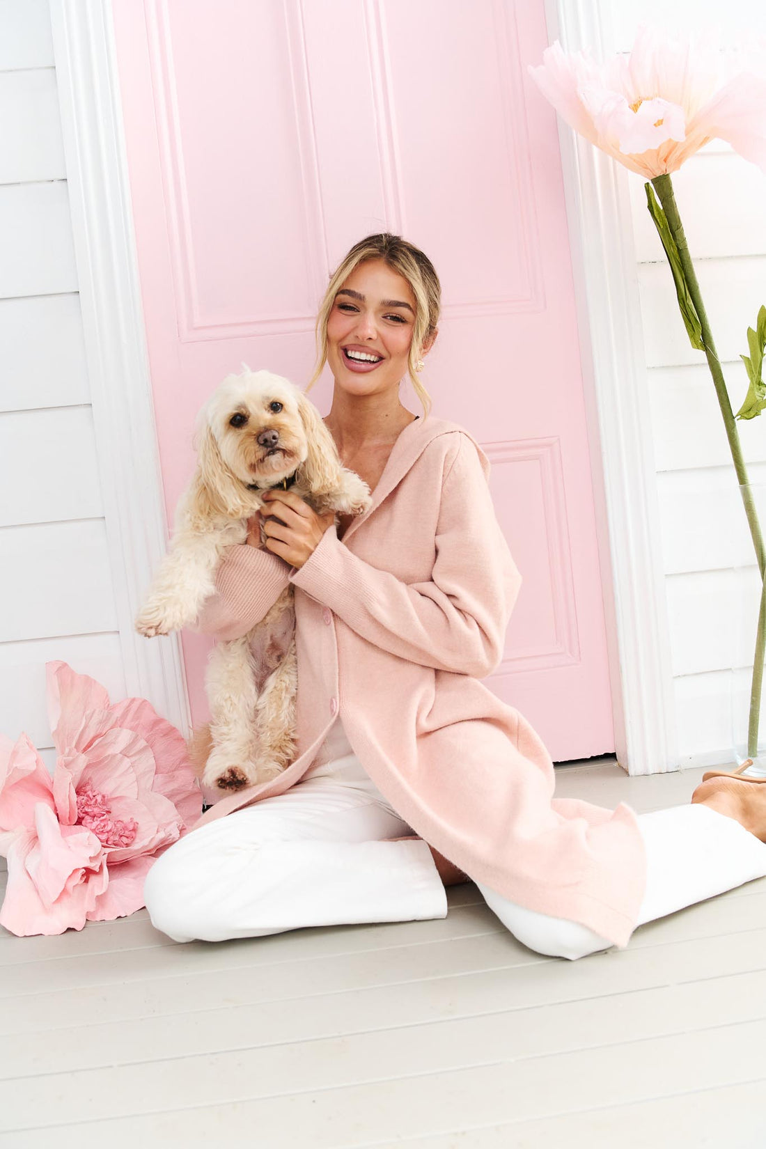 A woman sitting on a grey floor in front of a pink door, holding a cream-coloured dog. She is wearing the minnie + MAXXIE light pink Lennox hooded cardigan and white jeans.
