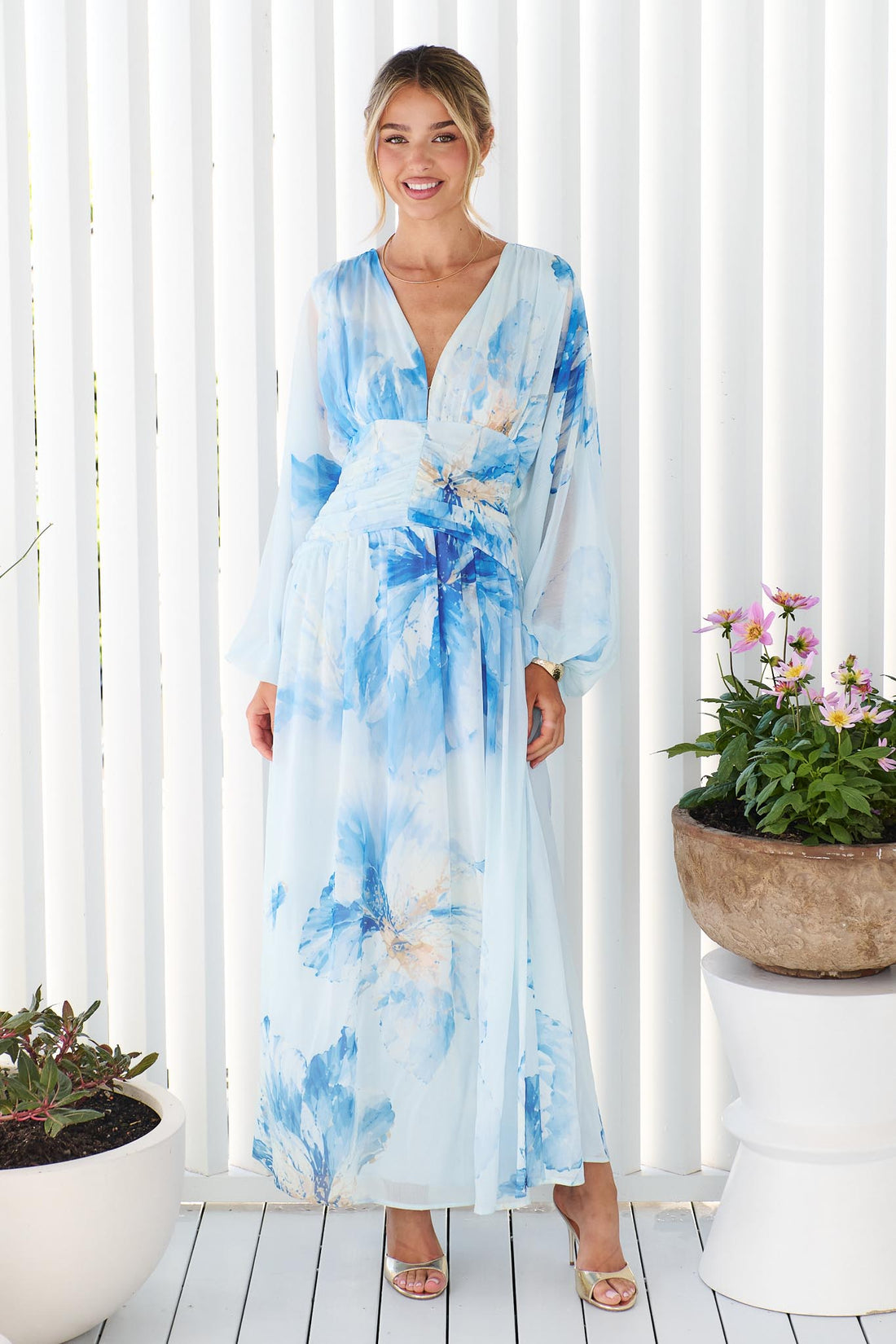 A woman wearing the Lola long-sleeve, V-neck maxi dress with a blue watercolor floral print, standing in front of a white slatted wall with potted plants on either side.