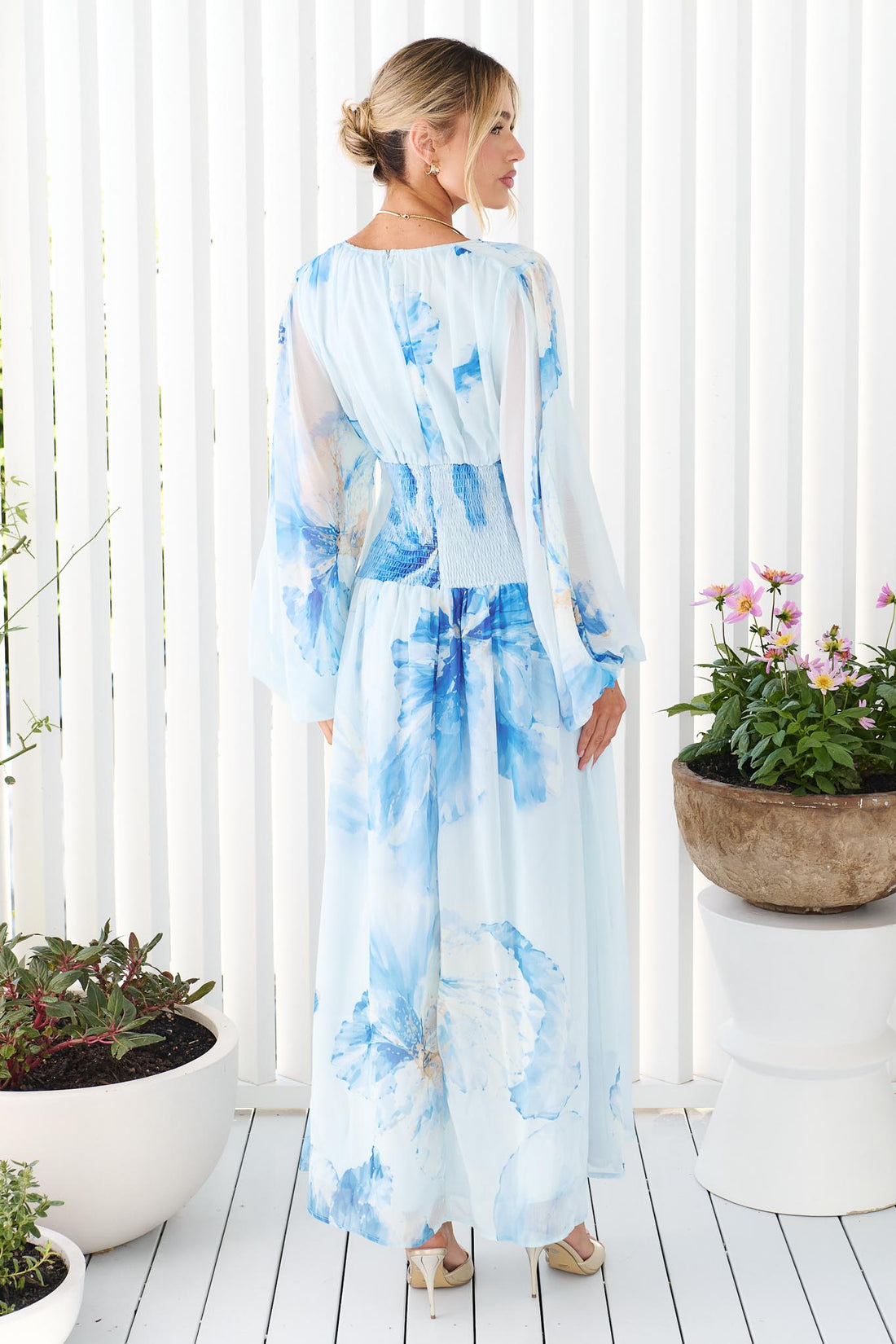 Back view of a woman wearing the minnie + MAXXIE Lola long-sleeve v-neck maxi dress with a blue watercolor floral print, standing in front of a white slatted wall with potted plants on either side.