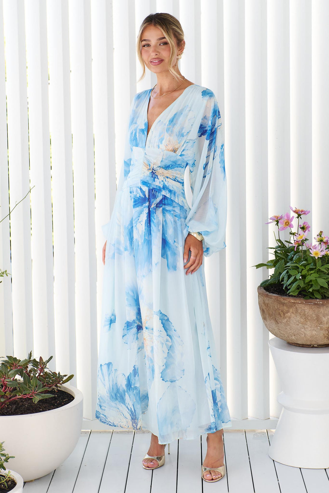 A woman wearing the Lola long-sleeve, V-neck maxi dress with a blue watercolor floral print, standing in front of a white slatted wall with potted plants on either side.