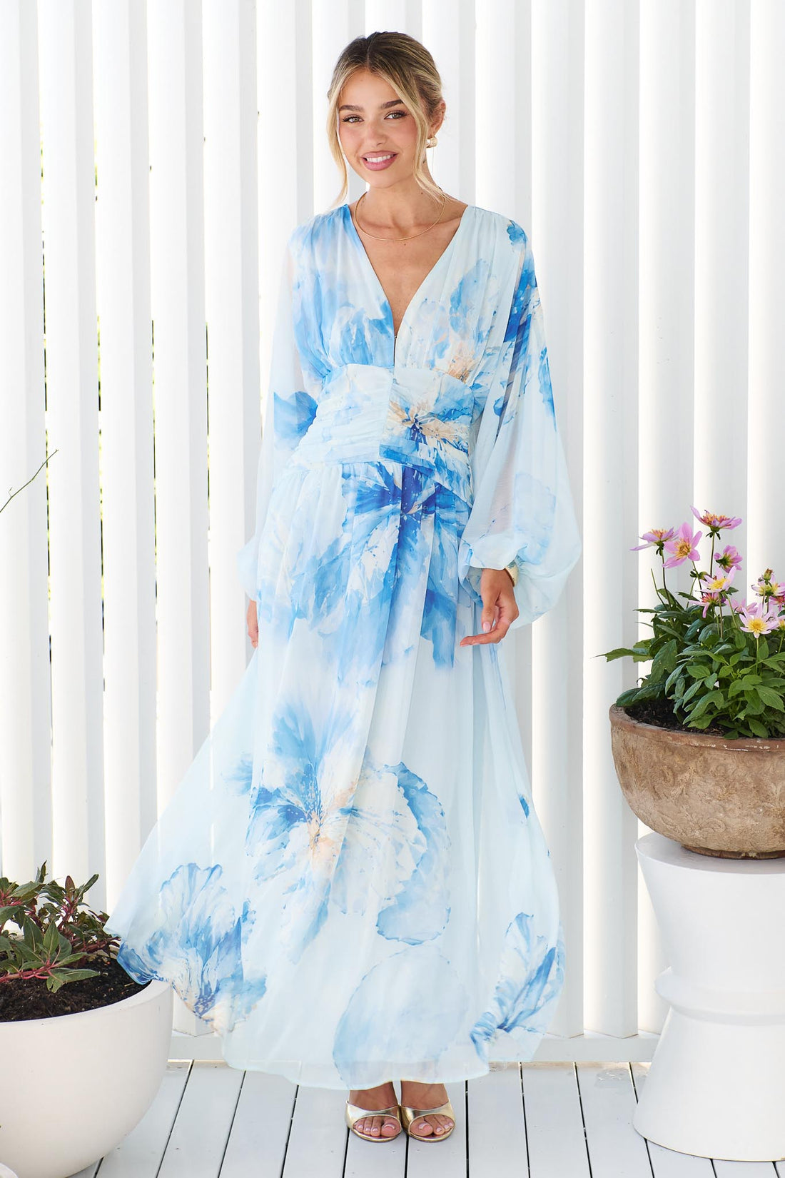 A woman wearing the minnie + MAXXIE Lola long-sleeve v-neck maxi dress with a blue watercolor floral print, standing in front of a white slatted wall with potted plants on either side.