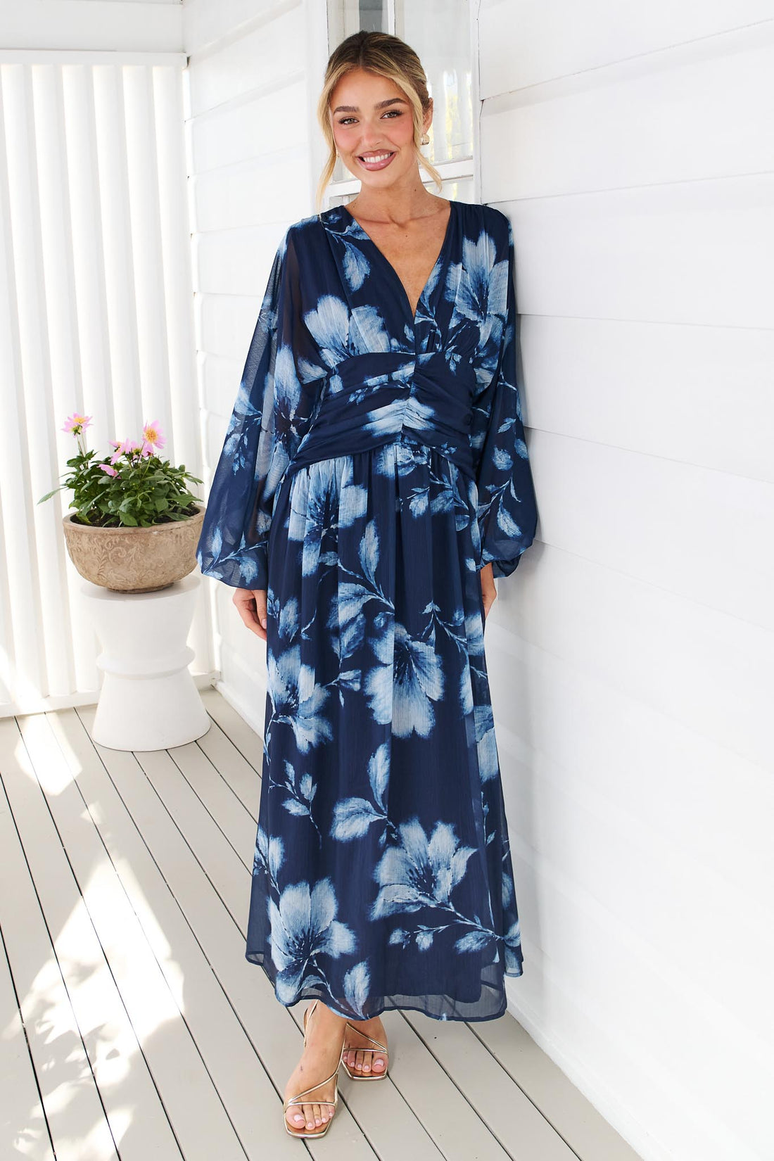 A woman wearing the minnie + MAXXIE Lola long-sleeve maxi dress in navy blue with a white floral print, standing against a white wall.