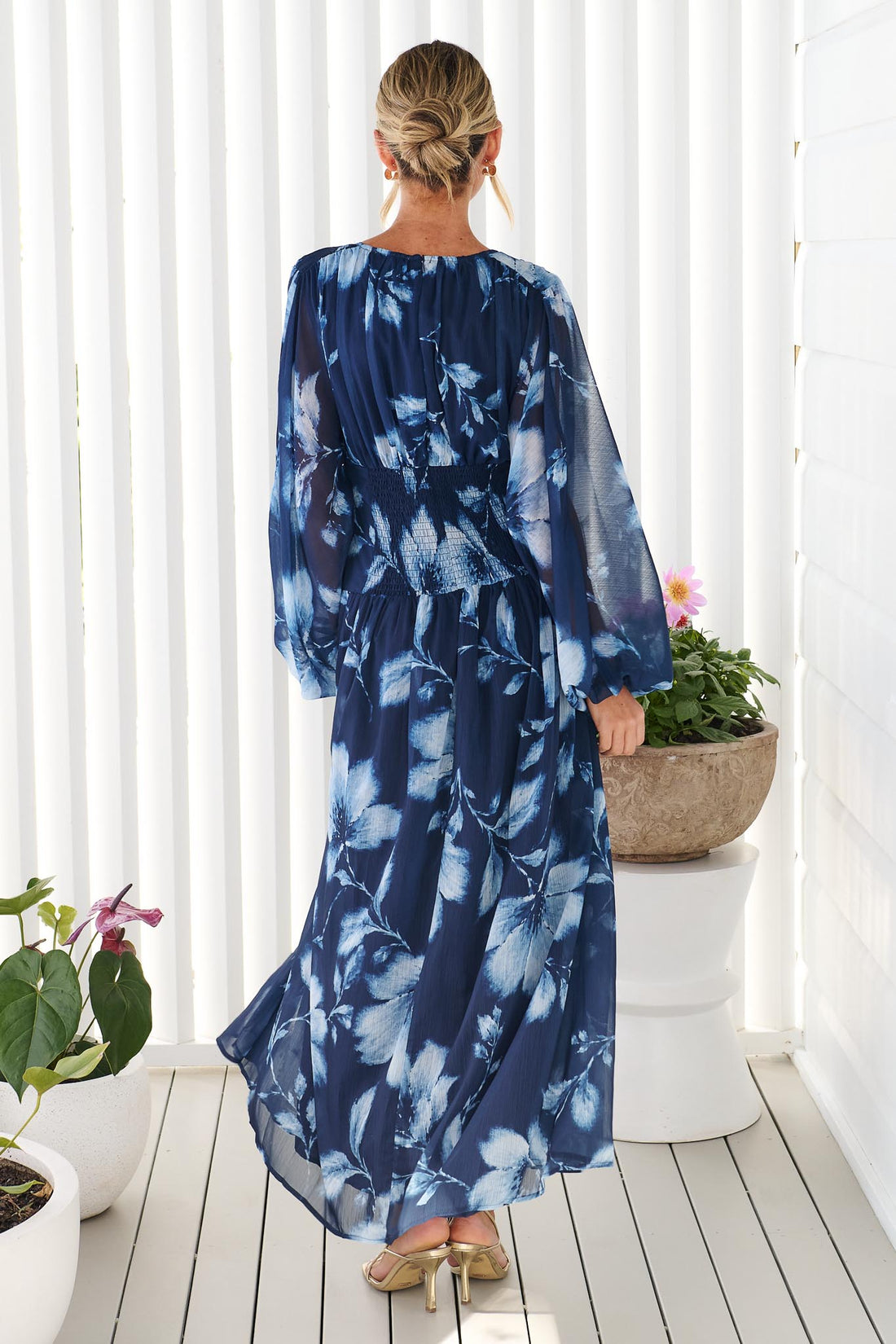 Back view of a woman wearing the minnie + MAXXIE Lola long-sleeve maxi dress in navy blue with a white floral print, standing against a white wall.