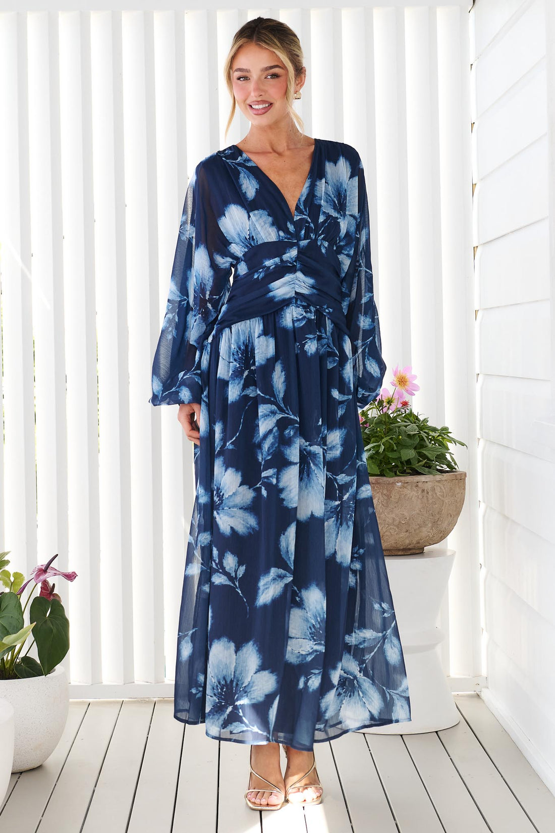 A woman wearing the minnie + MAXXIE Lola long-sleeve maxi dress in navy blue with a white floral print, standing against a white wall.