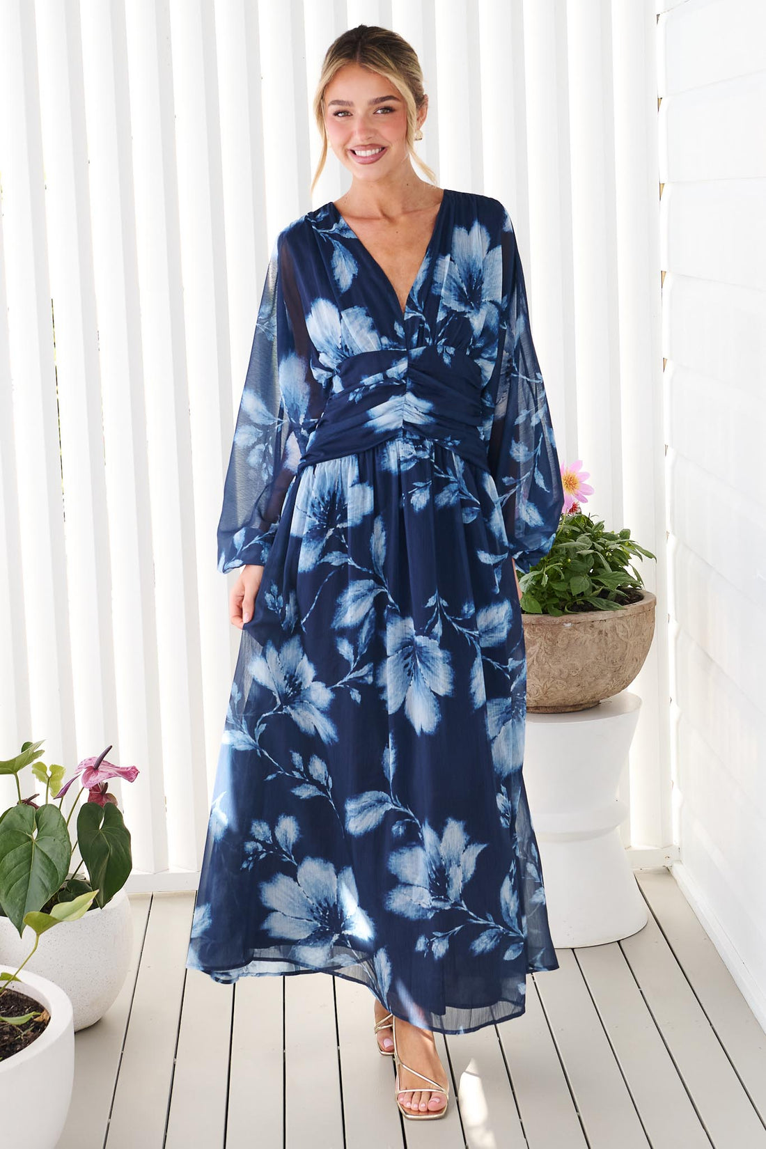 A woman wearing the minnie + MAXXIE Lola long-sleeve maxi dress in navy blue with a white floral print, standing against a white wall.