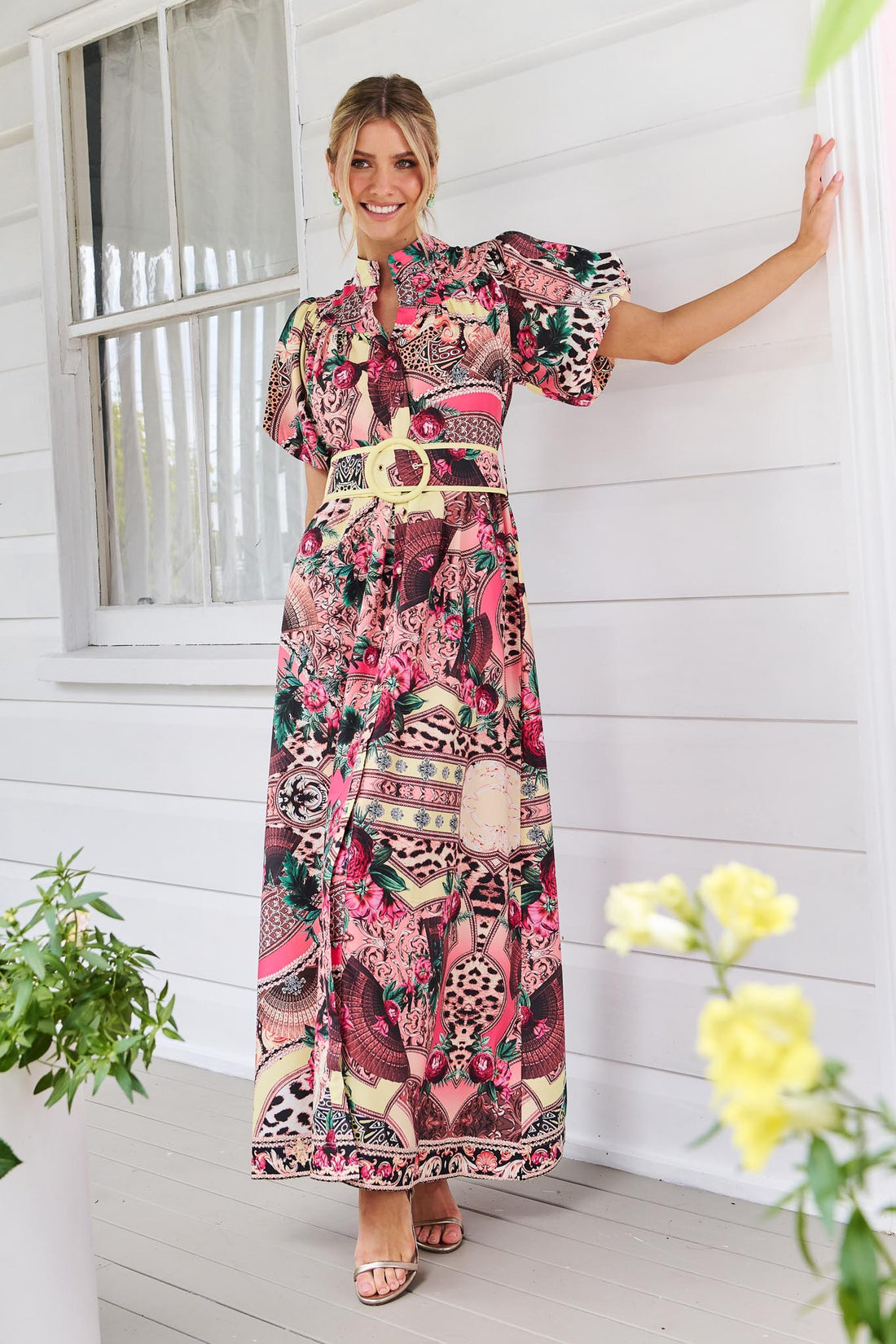 A woman wearing the minnie + MAXXIE Luisa maxi event dress with short puff-sleeves and a multicolour floral and abstract print, featuring a matching belt, standing against a white panelled wall.