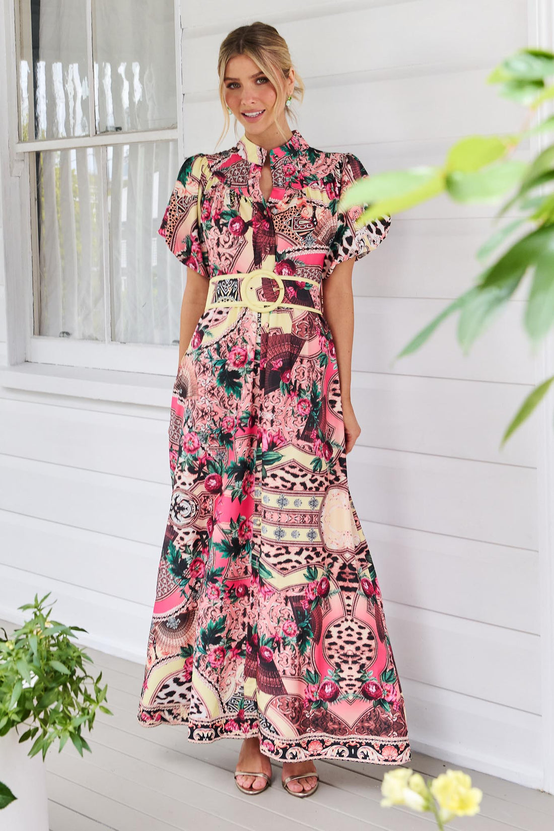 A woman wearing the minnie + MAXXIE Luisa maxi event dress with short puff-sleeves and a multicolour floral and abstract print, featuring a matching belt, standing against a white panelled wall.