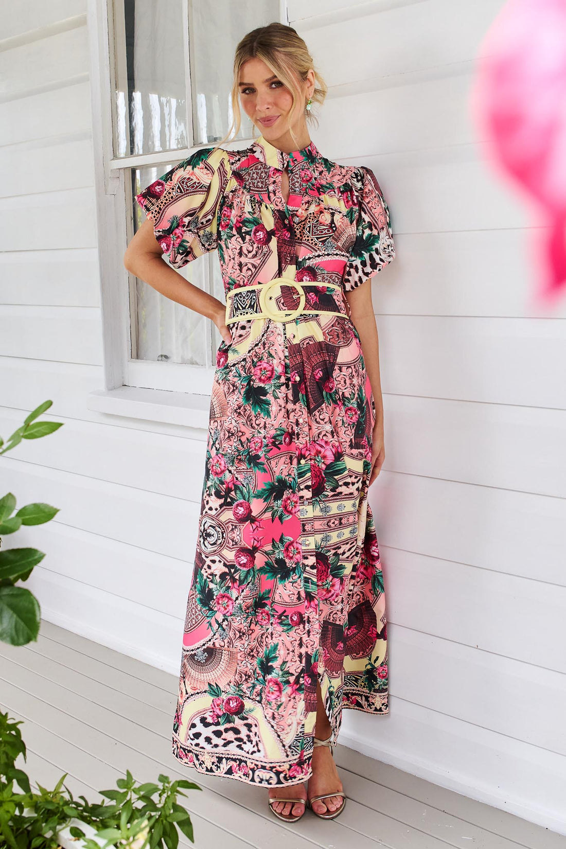 A woman wearing the minnie + MAXXIE Luisa maxi event dress with short puff-sleeves and a multicolour floral and abstract print, featuring a matching belt, standing against a white panelled wall.
