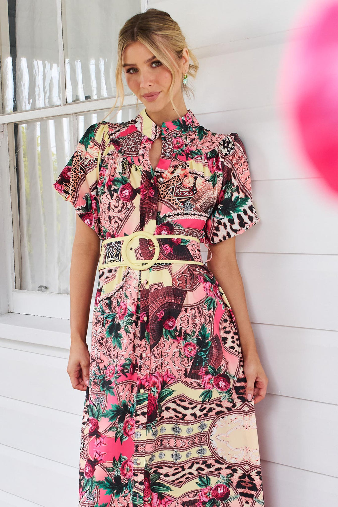 A woman wearing the minnie + MAXXIE Luisa maxi event dress with short puff-sleeves and a multicolour floral and abstract print, featuring a matching belt, standing against a white panelled wall.