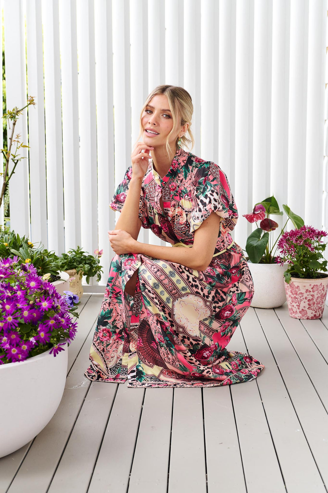 A woman wearing the minnie + MAXXIE Luisa maxi event dress with short puff-sleeves and a multicolour floral and abstract print, surrounded by potted flowers.