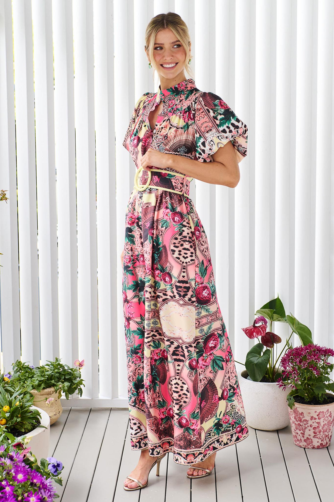 Side view of a woman wearing the minnie + MAXXIE Luisa maxi event dress with short puff-sleeves and a multicolour floral and abstract print, featuring a matching belt, standing against a white panelled wall.