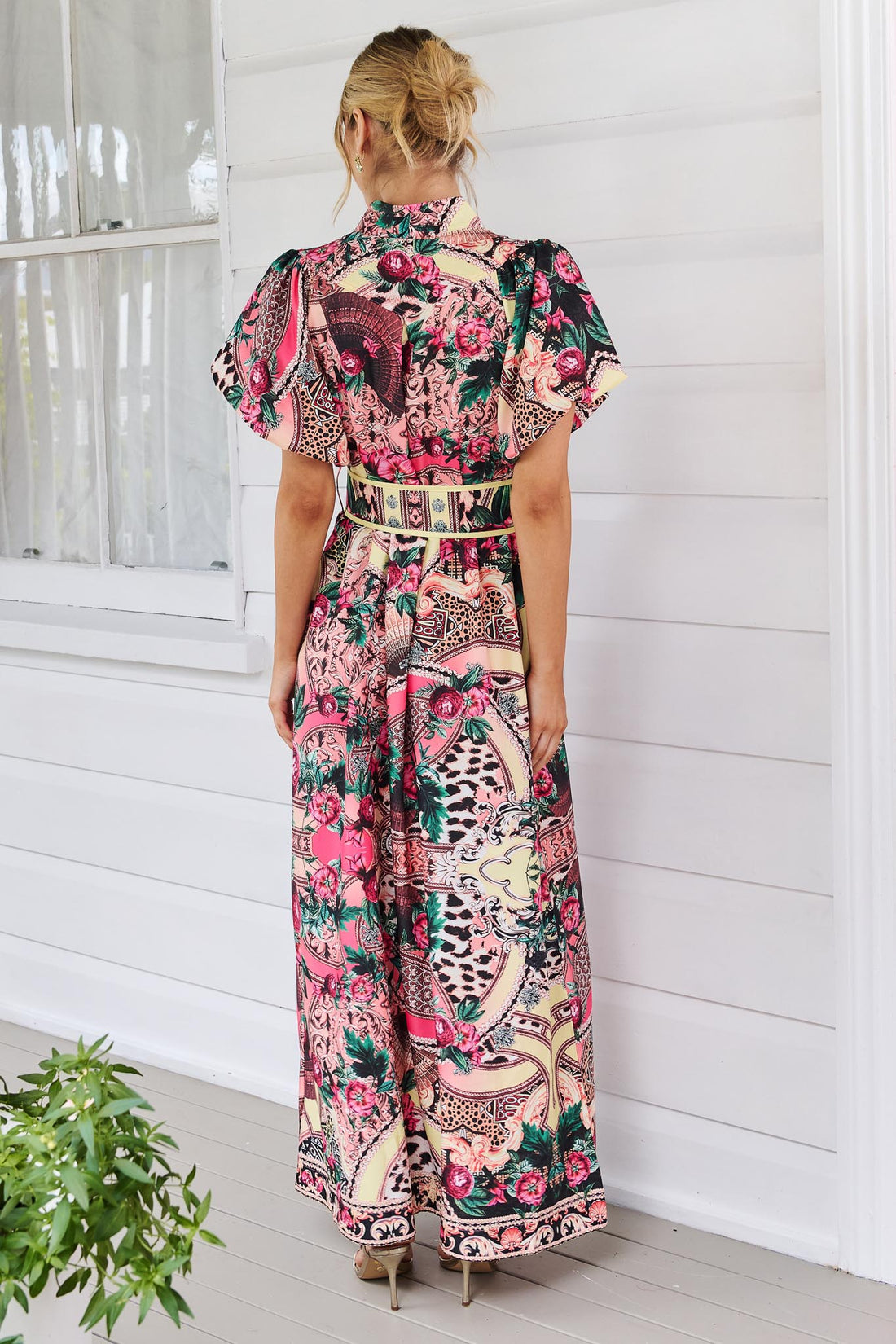 Back view of a woman wearing the minnie + MAXXIE Luisa maxi event dress with short puff-sleeves and a multicolour floral and abstract print, featuring a matching belt, standing against a white panelled wall.
