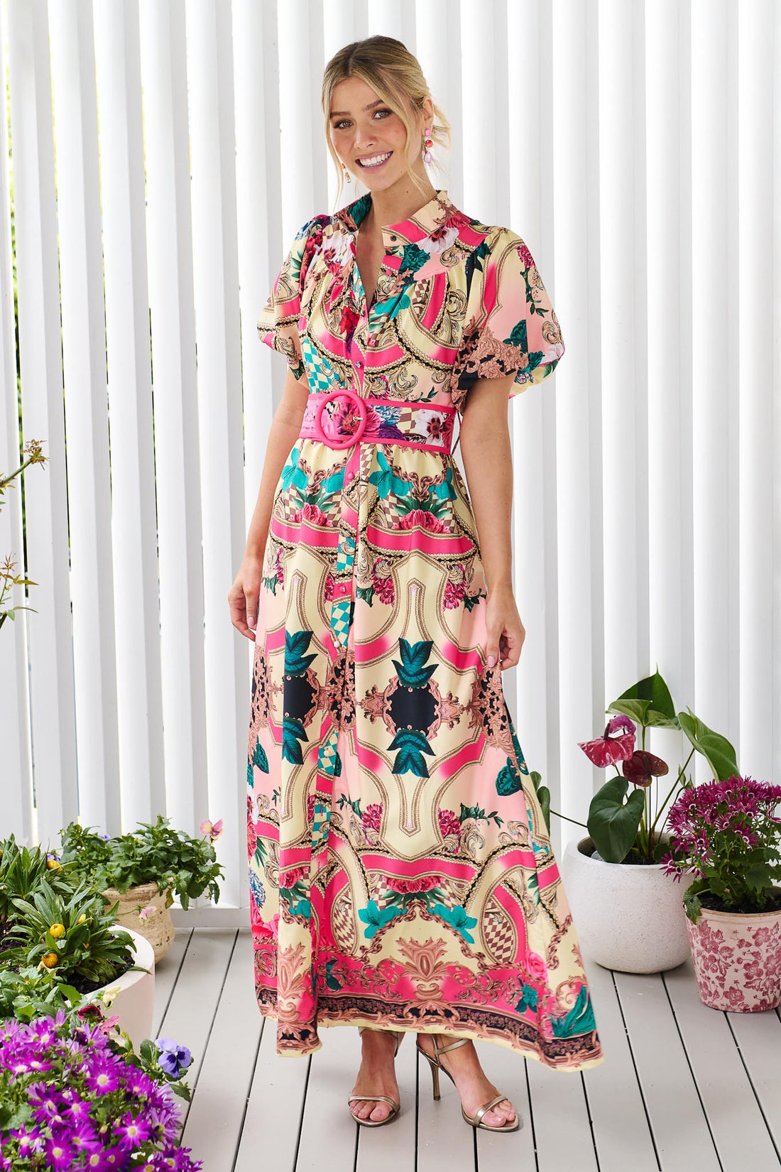 A woman wearing the minnie + MAXXIE Luisa maxi event dress with short puff-sleeves and a multicolour floral and abstract print, featuring a matching belt, standing against a white panelled wall.