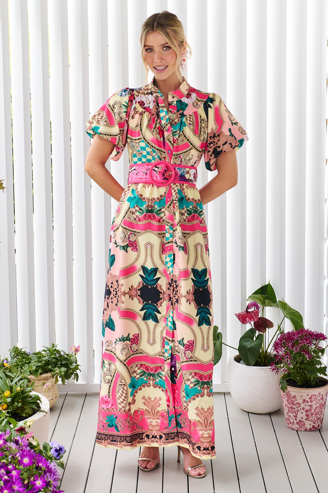 A woman wearing the minnie + MAXXIE Luisa maxi event dress with short puff-sleeves and a multicolour floral and abstract print, featuring a matching belt, standing against a white panelled wall.