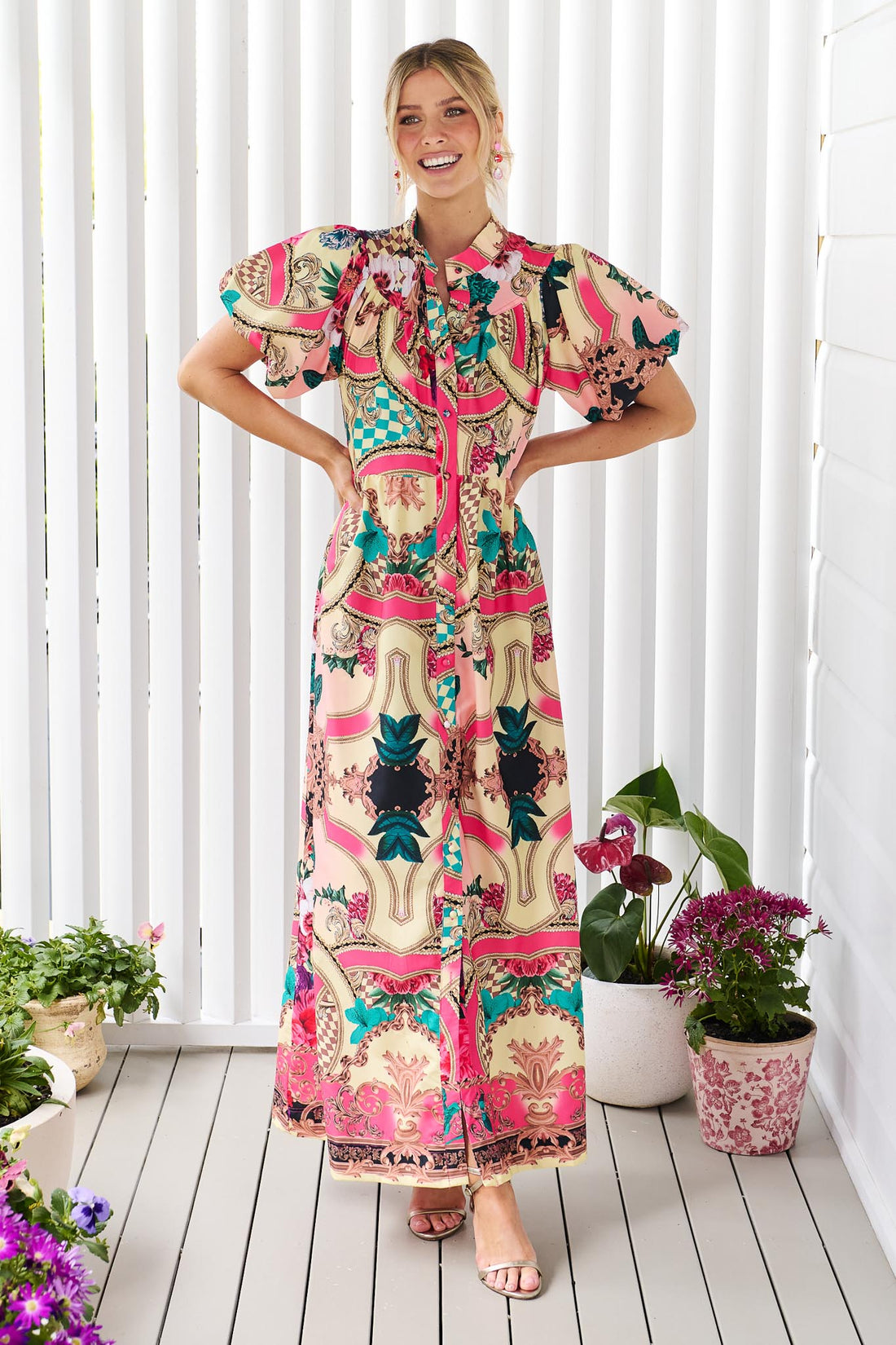 A woman wearing the minnie + MAXXIE Luisa maxi event dress with short puff-sleeves and a multicolour floral and abstract print, standing against a white panelled wall.