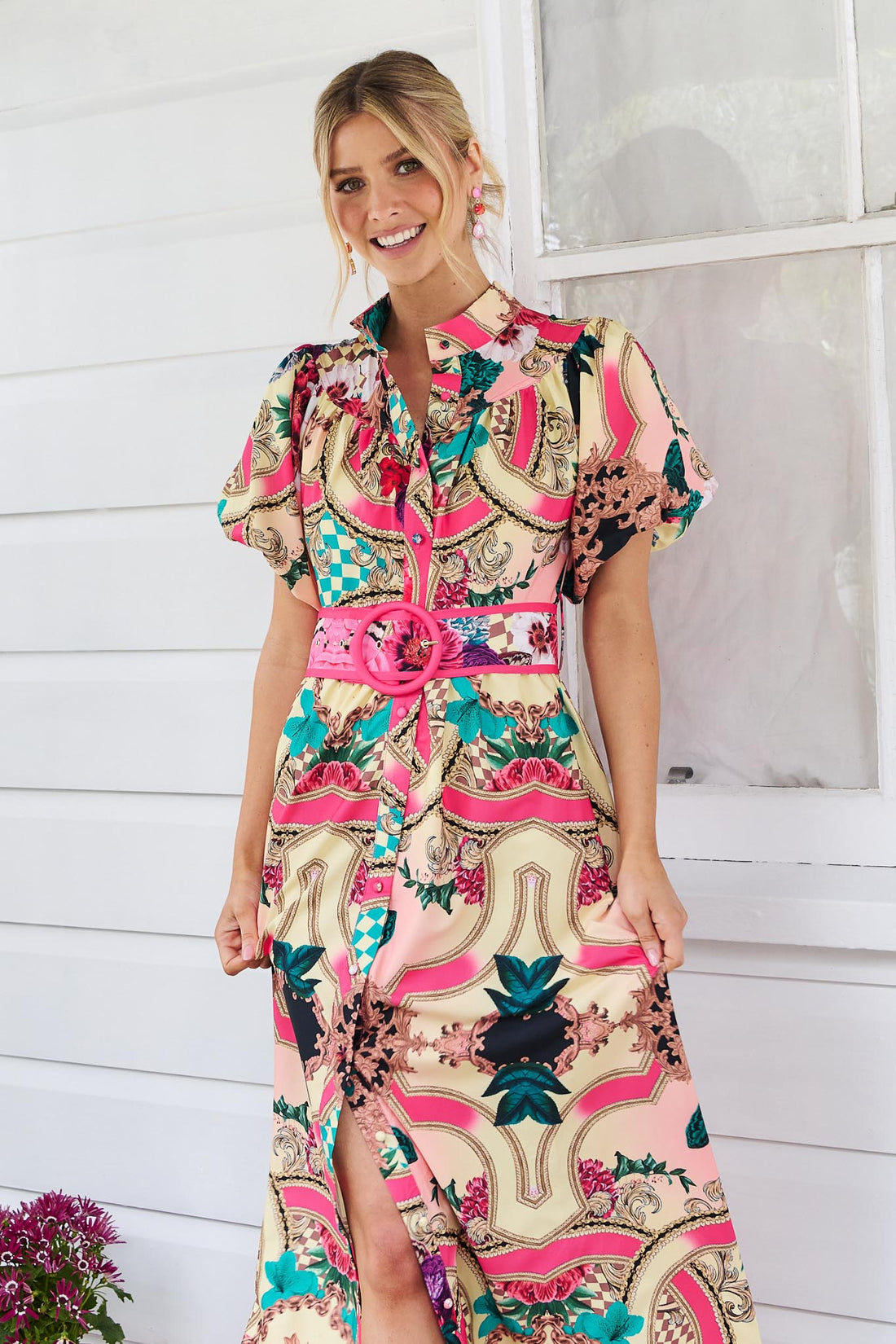 A woman wearing the minnie + MAXXIE Luisa maxi event dress with short puff-sleeves and a multicolour floral and abstract print, featuring a matching belt, standing against a white panelled wall.