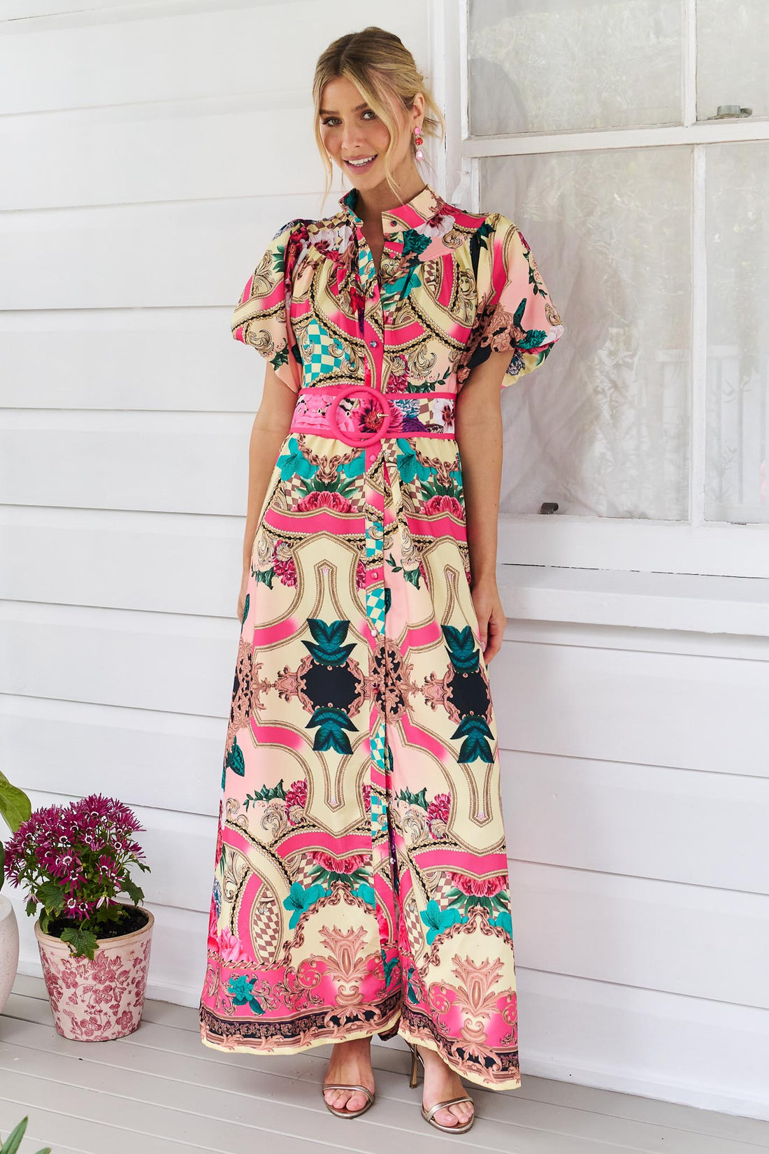 A woman wearing the minnie + MAXXIE Luisa maxi event dress with short puff-sleeves and a multicolour floral and abstract print, featuring a matching belt, standing against a white panelled wall.