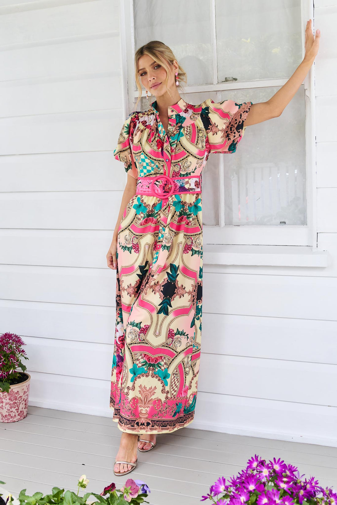A woman wearing the minnie + MAXXIE Luisa maxi event dress with short puff-sleeves and a multicolour floral and abstract print, featuring a matching belt, standing against a white panelled wall.