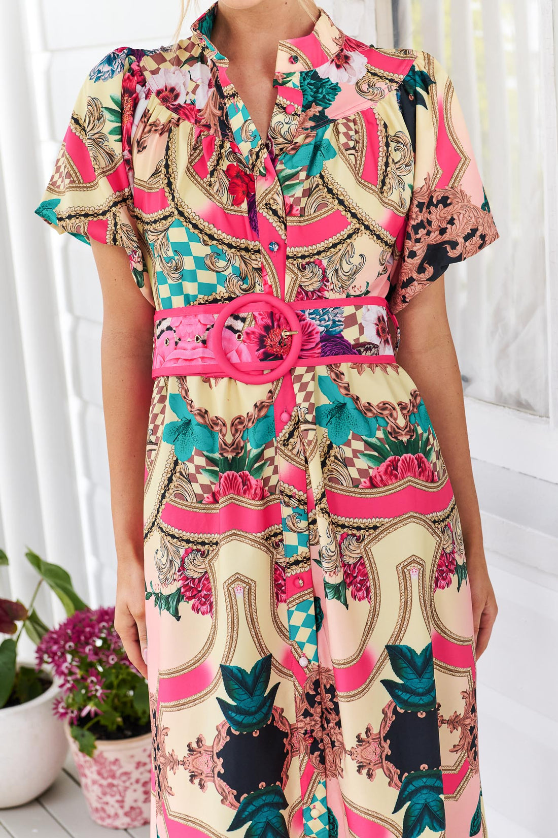 Detail of a woman wearing the minnie + MAXXIE Luisa maxi event dress with short puff-sleeves and a multicolour floral and abstract print, featuring a matching belt, standing against a white panelled wall.