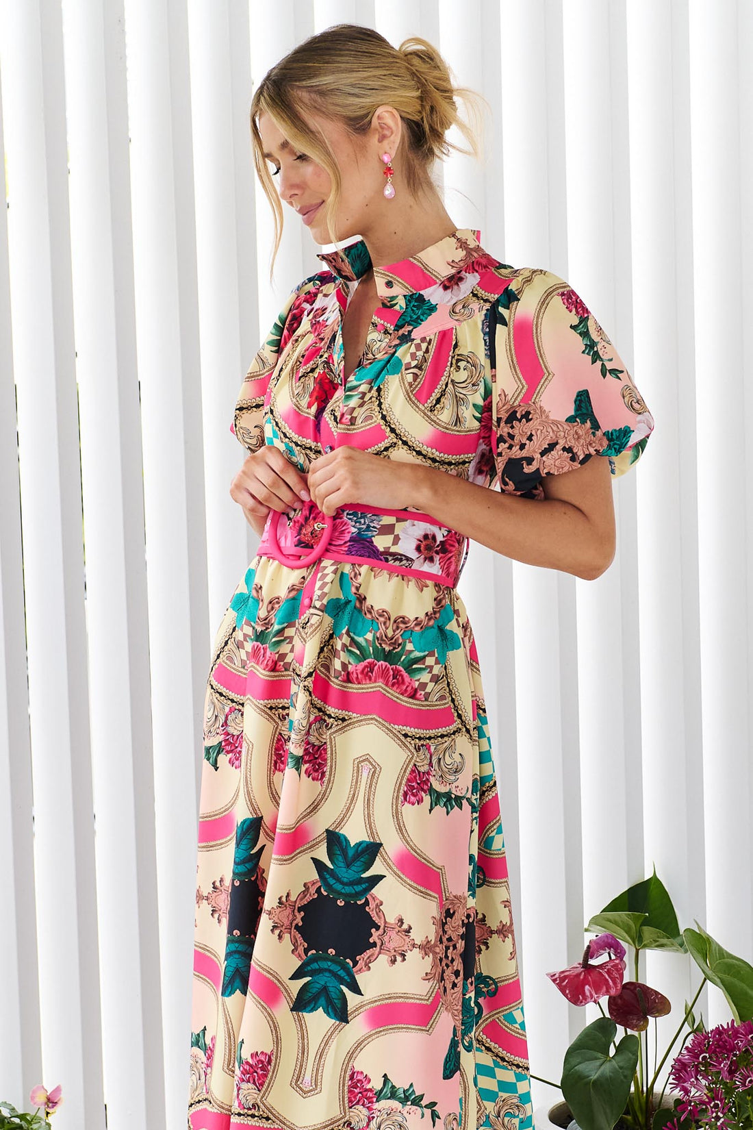 A woman wearing the minnie + MAXXIE Luisa maxi event dress with short puff-sleeves and a multicolour floral and abstract print, featuring a matching belt, standing against a white panelled wall.