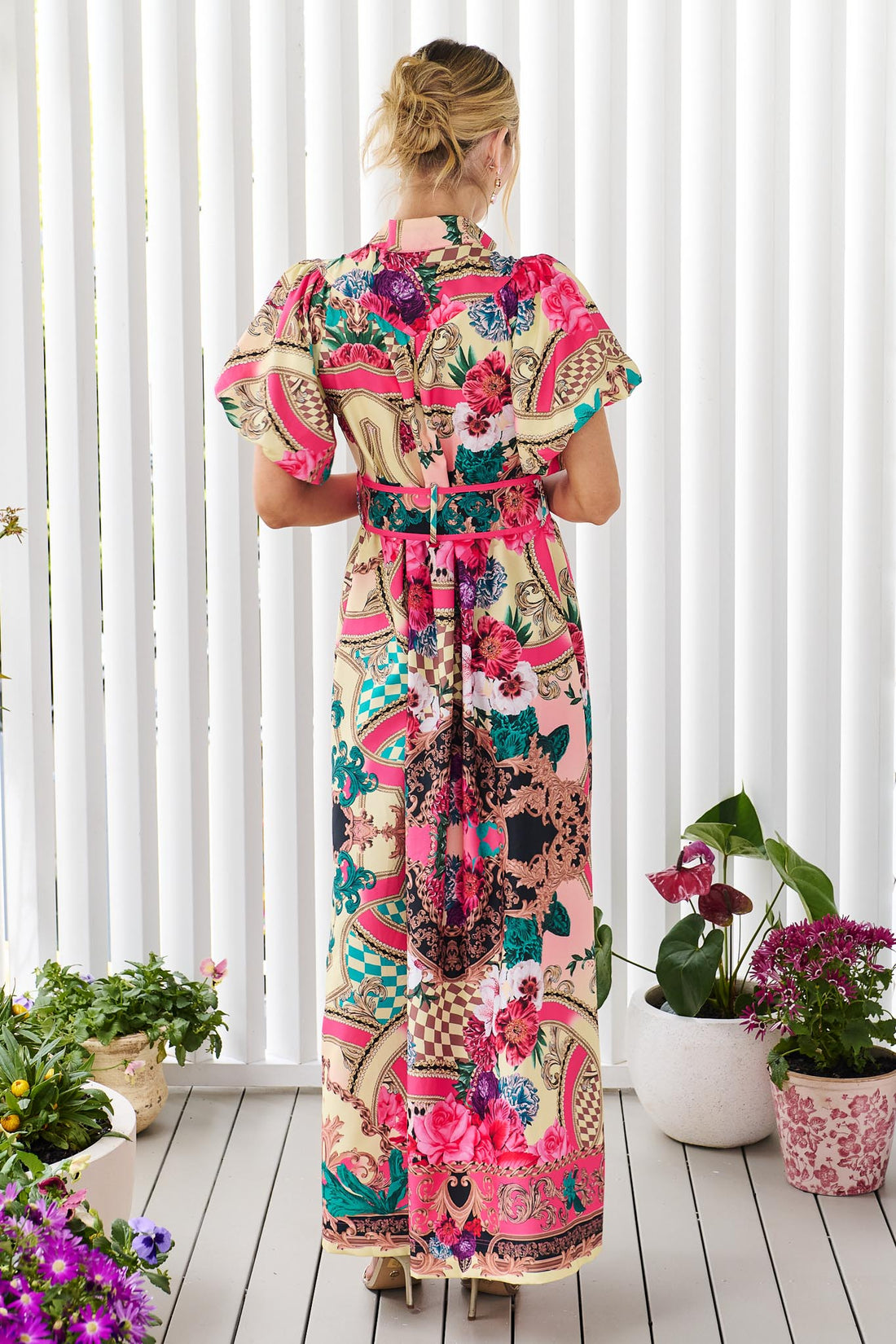 Back view of a woman wearing the minnie + MAXXIE Luisa maxi event dress with short puff-sleeves and a multicolour floral and abstract print, featuring a matching belt, standing against a white panelled wall.