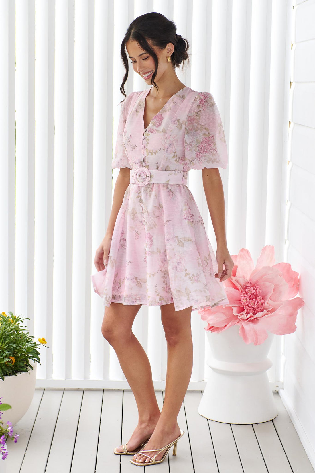 A woman wearing the minnie + MAXXIE Madeleine pink floral mini dress with short puff sleeves, a V-neckline, and a matching fabric belt with a round buckle, set against a white panelled wall.