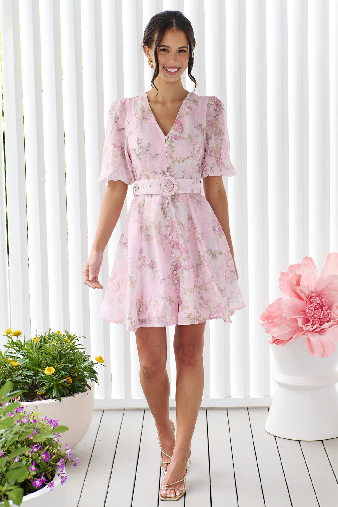A woman wearing the minnie + MAXXIE Madeleine pink floral mini dress with short puff sleeves, a V-neckline, and a matching fabric belt with a round buckle, set against a white panelled wall.