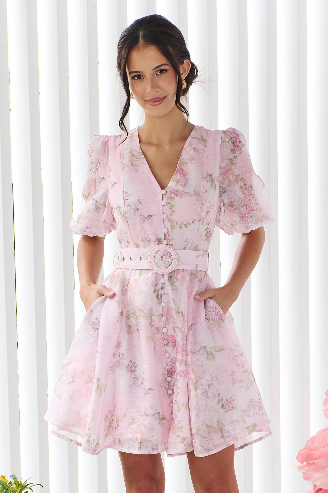 A woman wearing the minnie + MAXXIE Madeleine pink floral mini dress with short puff sleeves, a V-neckline, and a matching fabric belt with a round buckle, set against a white panelled wall.