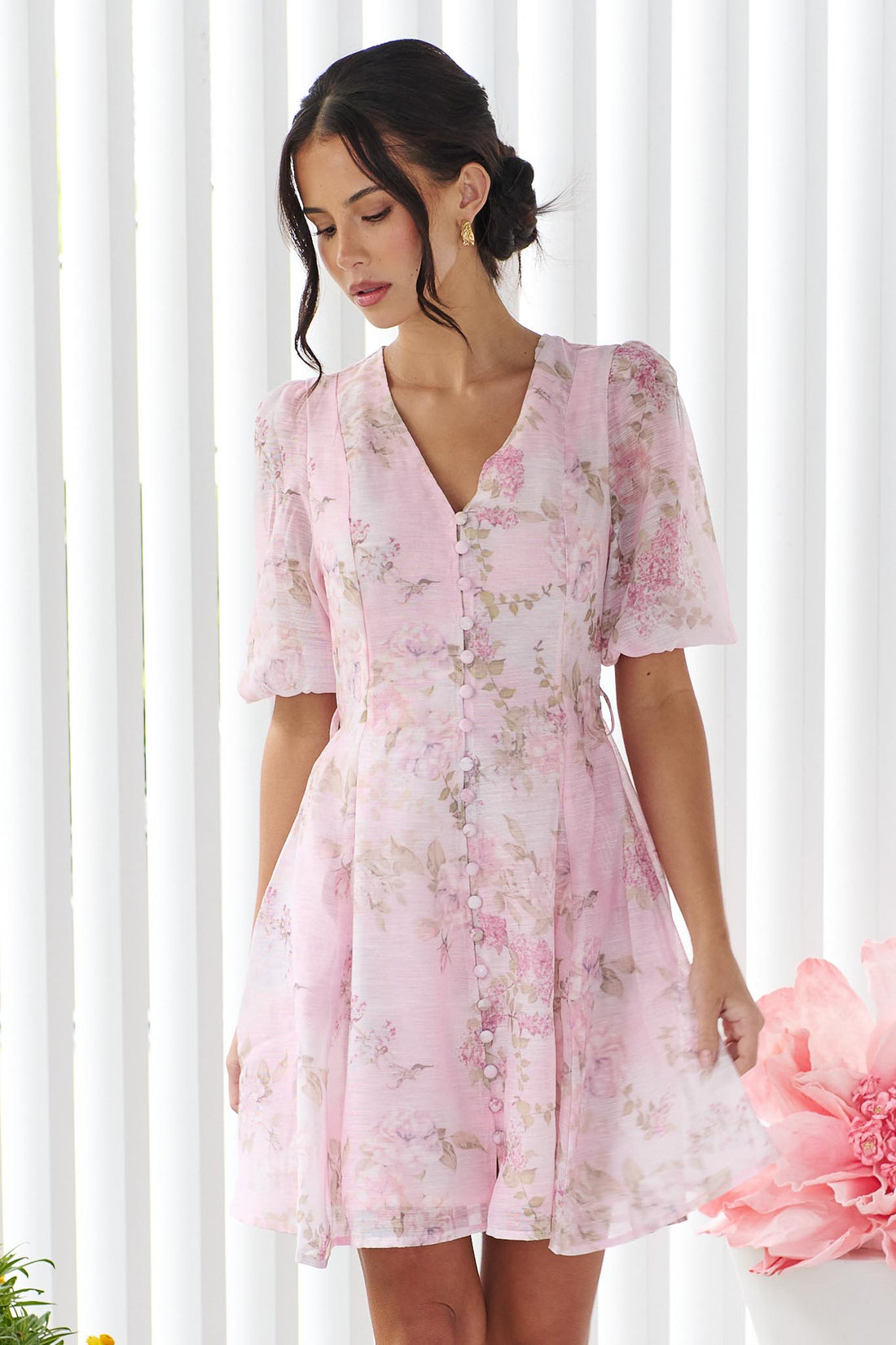 A woman wearing the minnie + MAXXIE Madeleine pink floral mini dress with short puff sleeves, and a V-neckline, set against a white panelled wall.