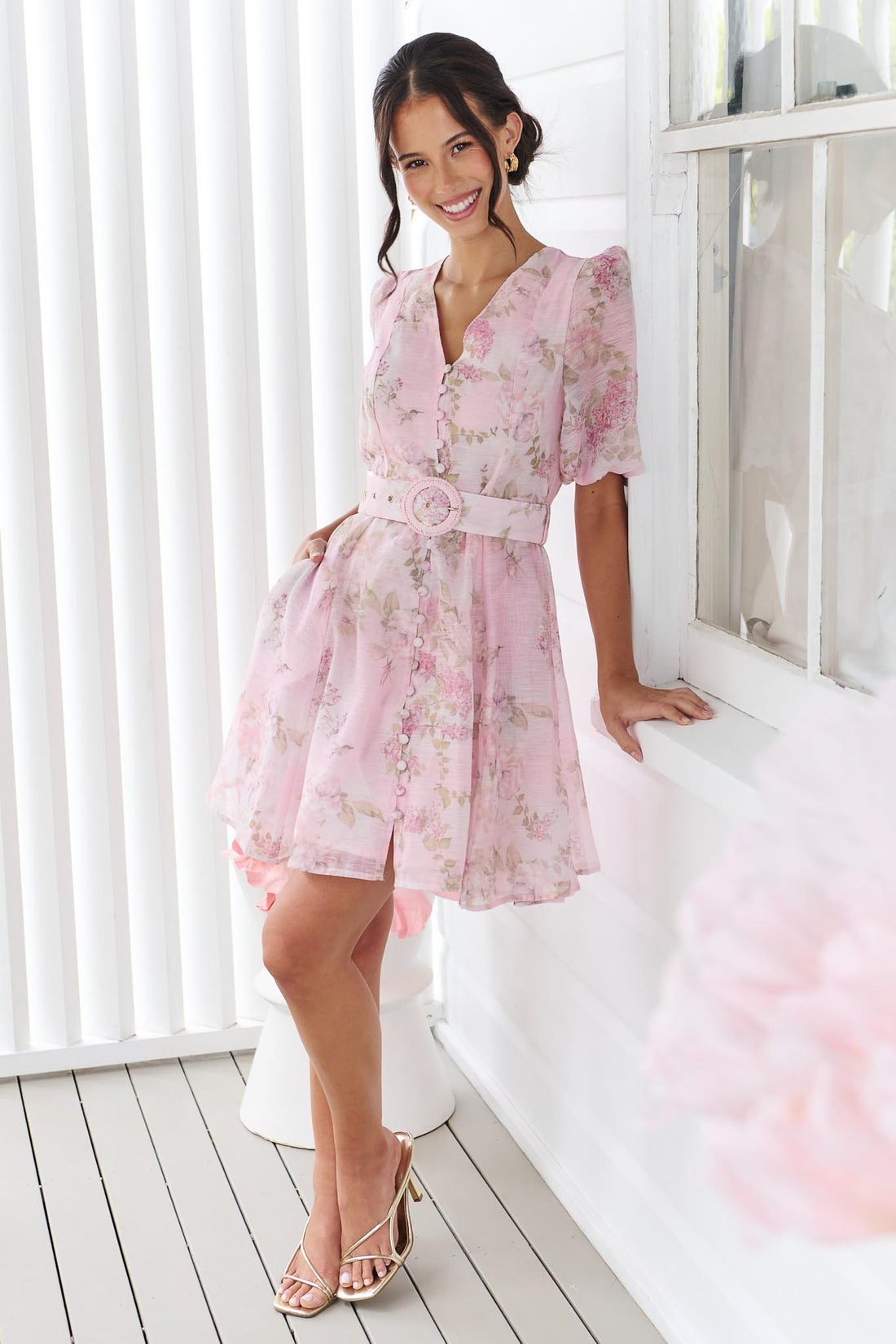 A woman wearing the minnie + MAXXIE Madeleine pink floral mini dress with short puff sleeves, a V-neckline, and a matching fabric belt with a round buckle, set against a white panelled wall.