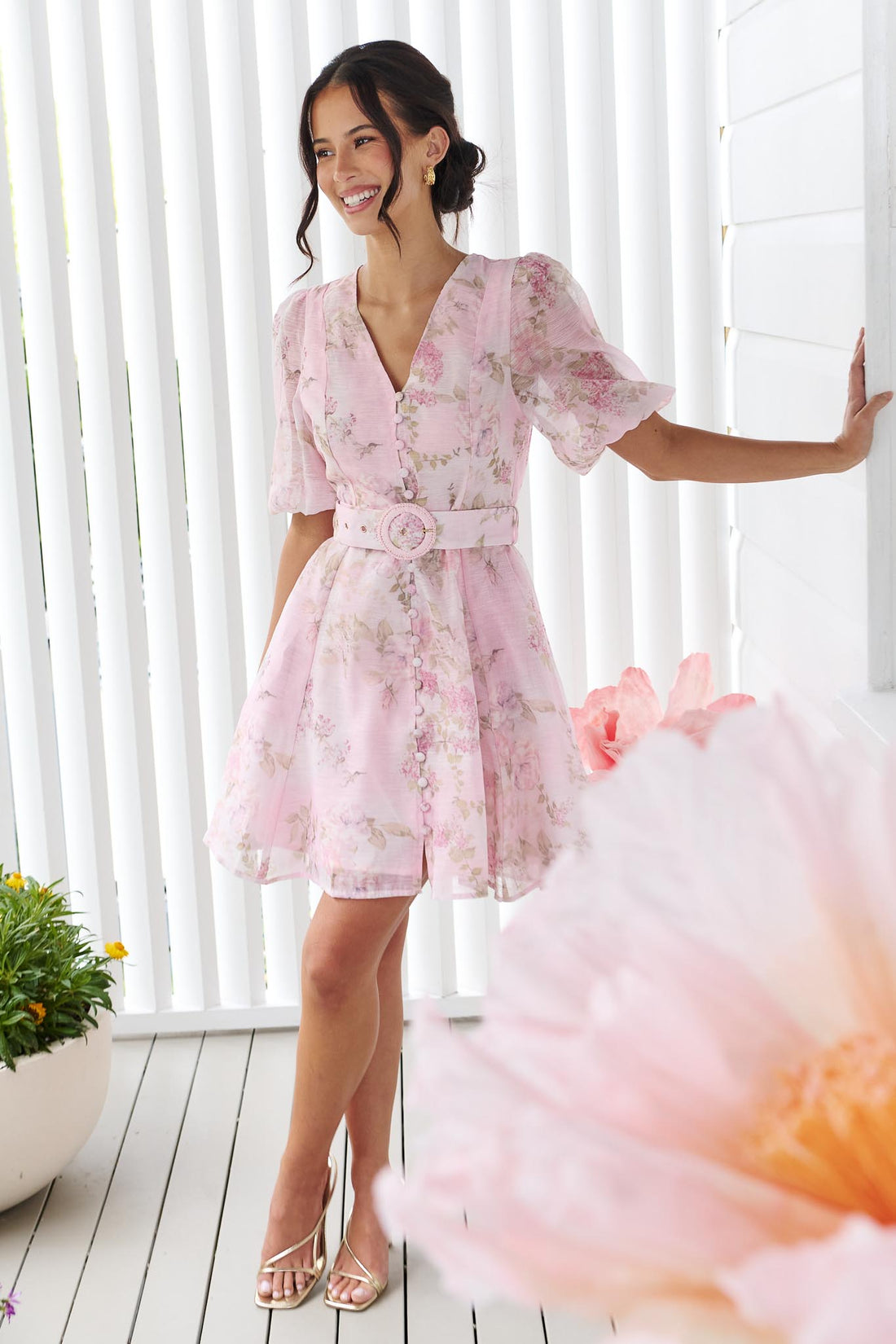 A woman wearing the minnie + MAXXIE Madeleine pink floral mini dress with short puff sleeves, a V-neckline, and a matching fabric belt with a round buckle, set against a white panelled wall.