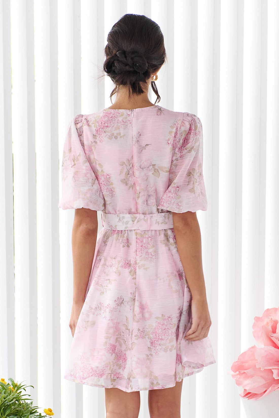 Back view of a woman wearing the minnie + MAXXIE Madeleine pink floral mini dress with short puff sleeves, a V-neckline, and a matching fabric belt with a round buckle, set against a white panelled wall.