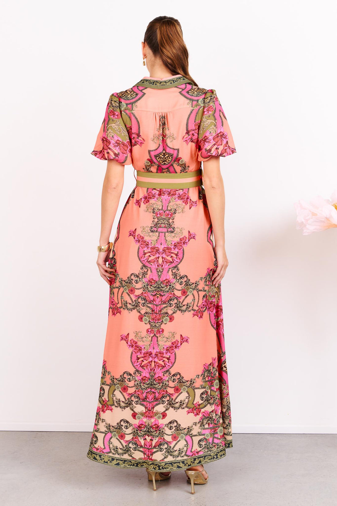 Back view of a woman wearing the minnie + MAXXIE Madison Garden peachy pink floral maxi event dress, standing against a white background.